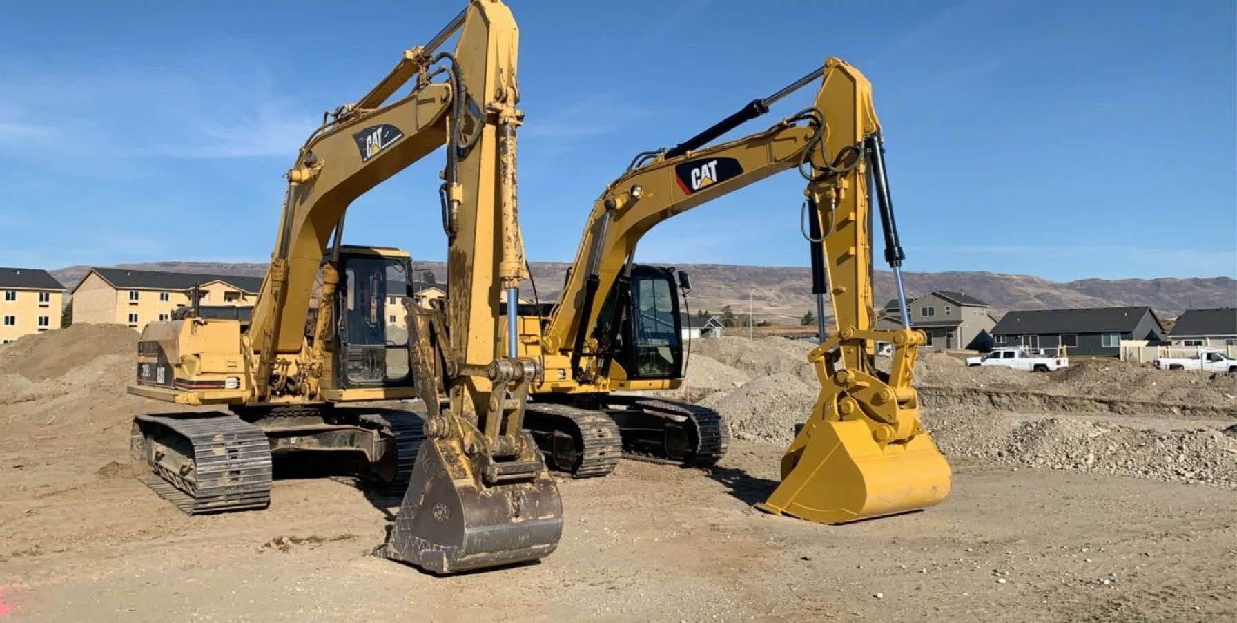 Two yellow excavators parked at a dusty construction site with houses in the background