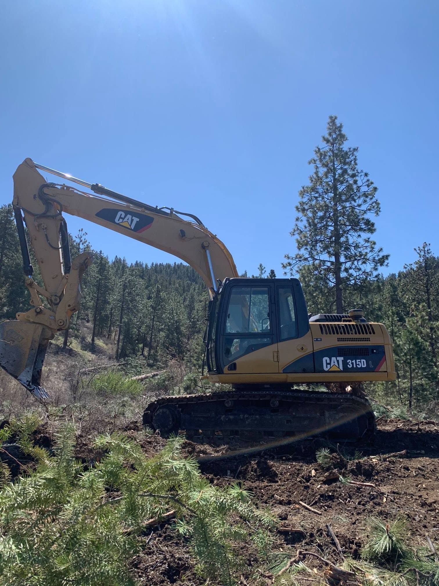 Yellow CAT excavator clearing brush on a wooded hillside under a clear blue sky