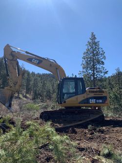 Yellow CAT excavator clearing brush on a wooded hillside under a clear blue sky