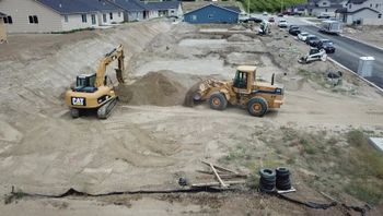 Two yellow construction machines grading a sandy lot near houses and a road