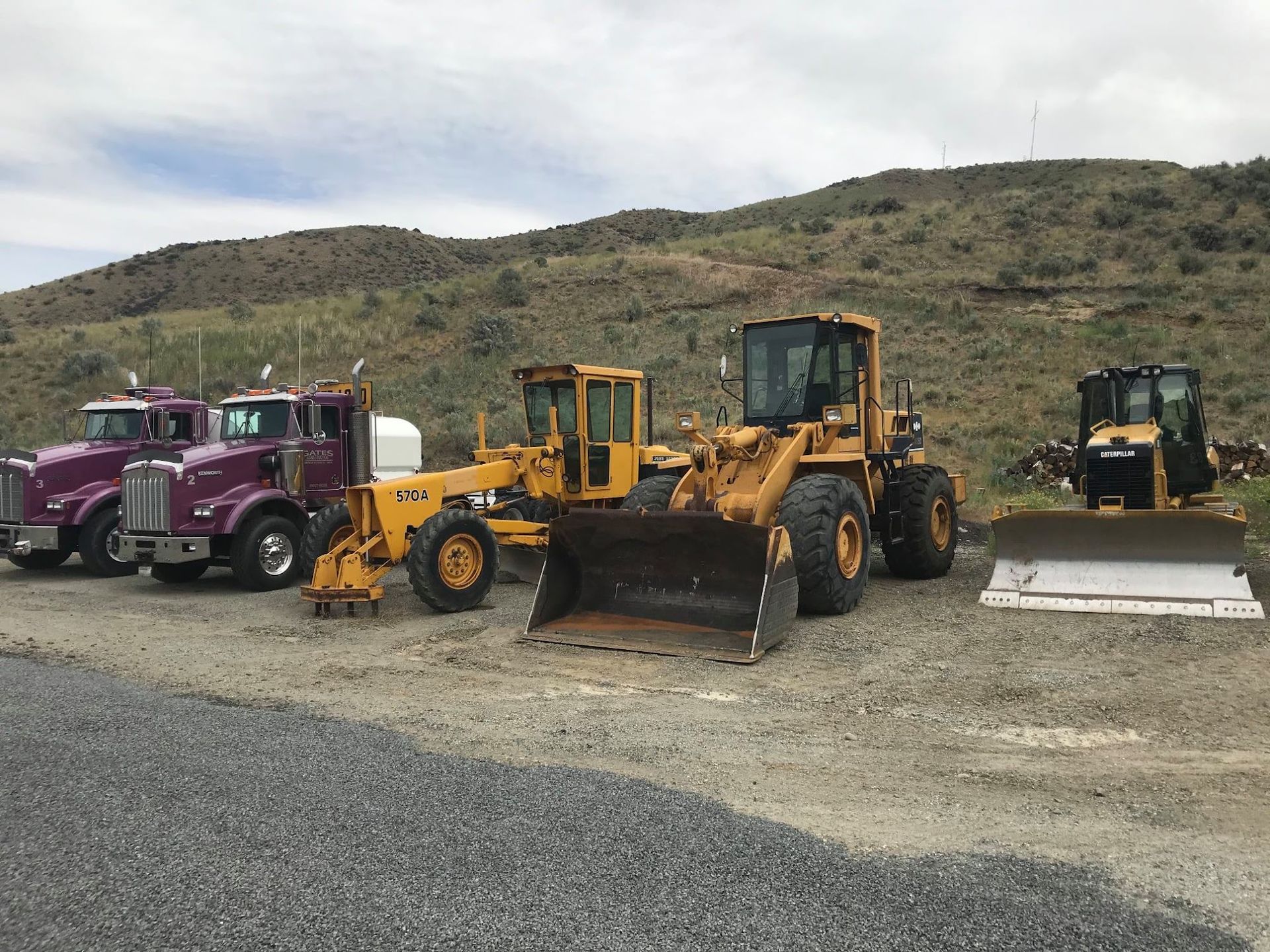 Construction vehicles lined up on gravel, including a purple truck and yellow loaders, with hills in the background