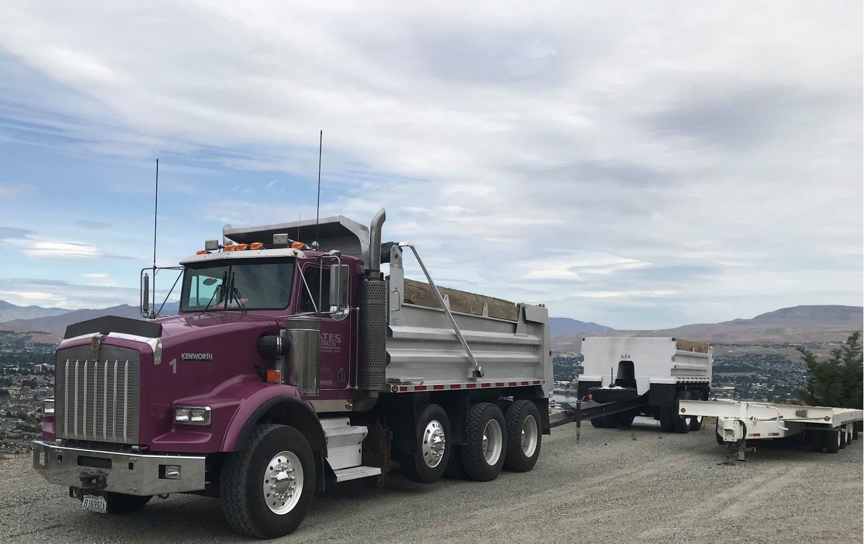 Purple semi truck towing a trailer on a roadside with hills in the background