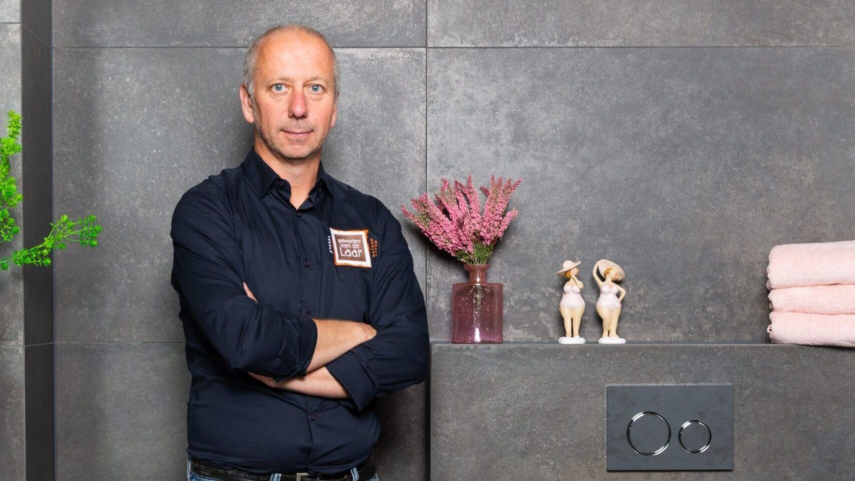 Een man met gekruiste armen staat bij een muur met decoratieve voorwerpen.