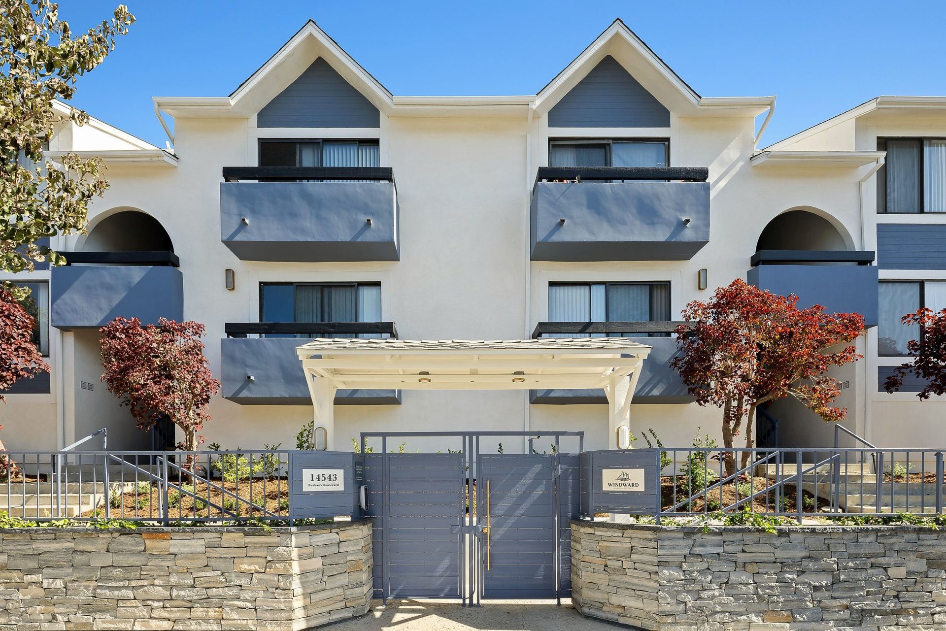 Modern apartment building entrance with a gated fence, balconies, and red-leafed trees