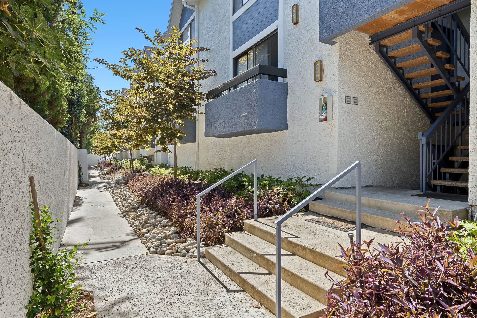Apartment stairway and walkway with metal handrails, gravel path, and landscaped shrubs beside a building.