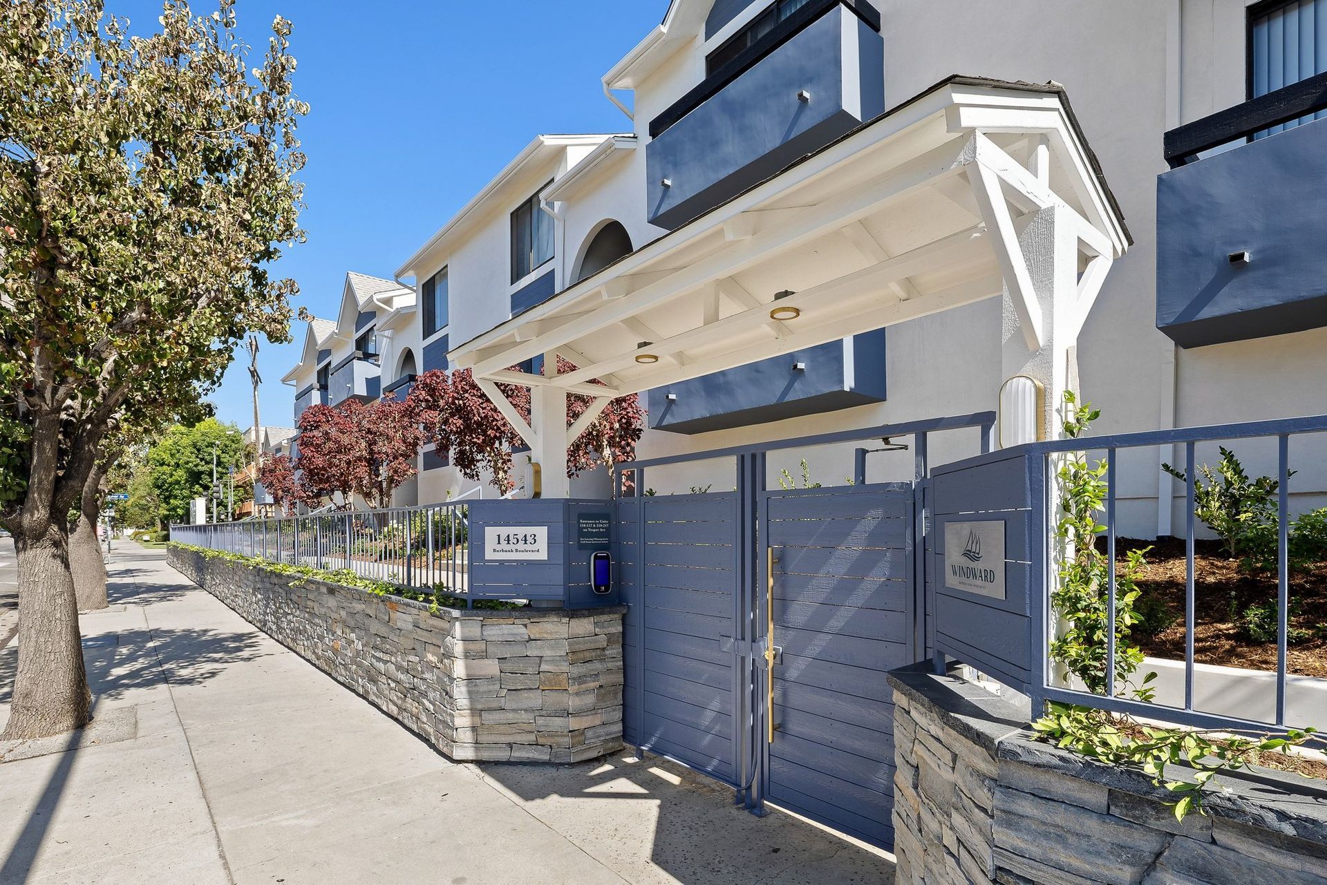 Modern apartment entrance with blue gate, stone pillars, and sidewalk along a tree-lined street