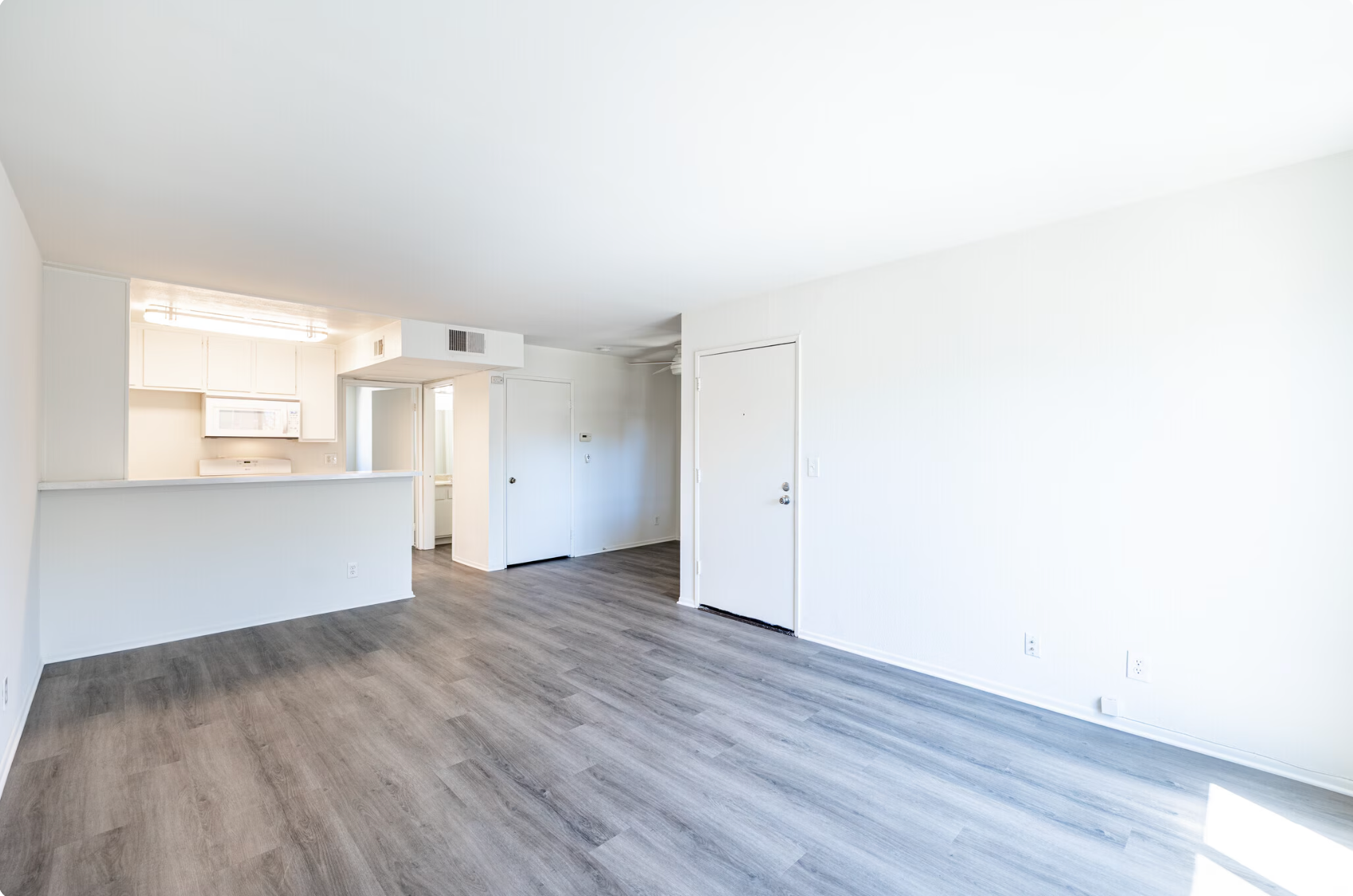 Empty bright living room with gray wood floors, open kitchen, and white walls