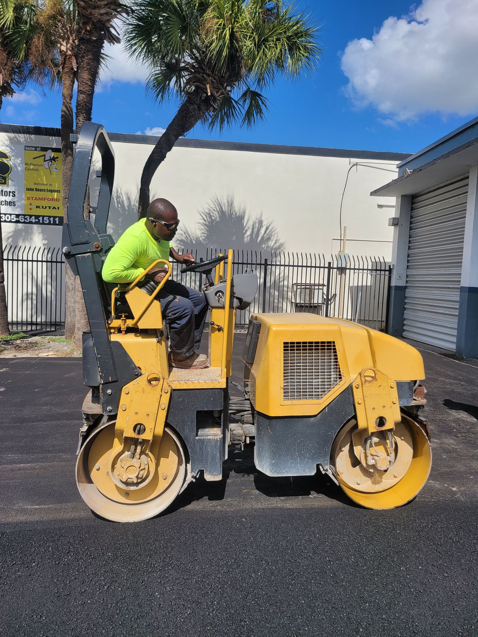 A person in a bright yellow safety shirt operates a yellow industrial road roller on an asphalt surface.