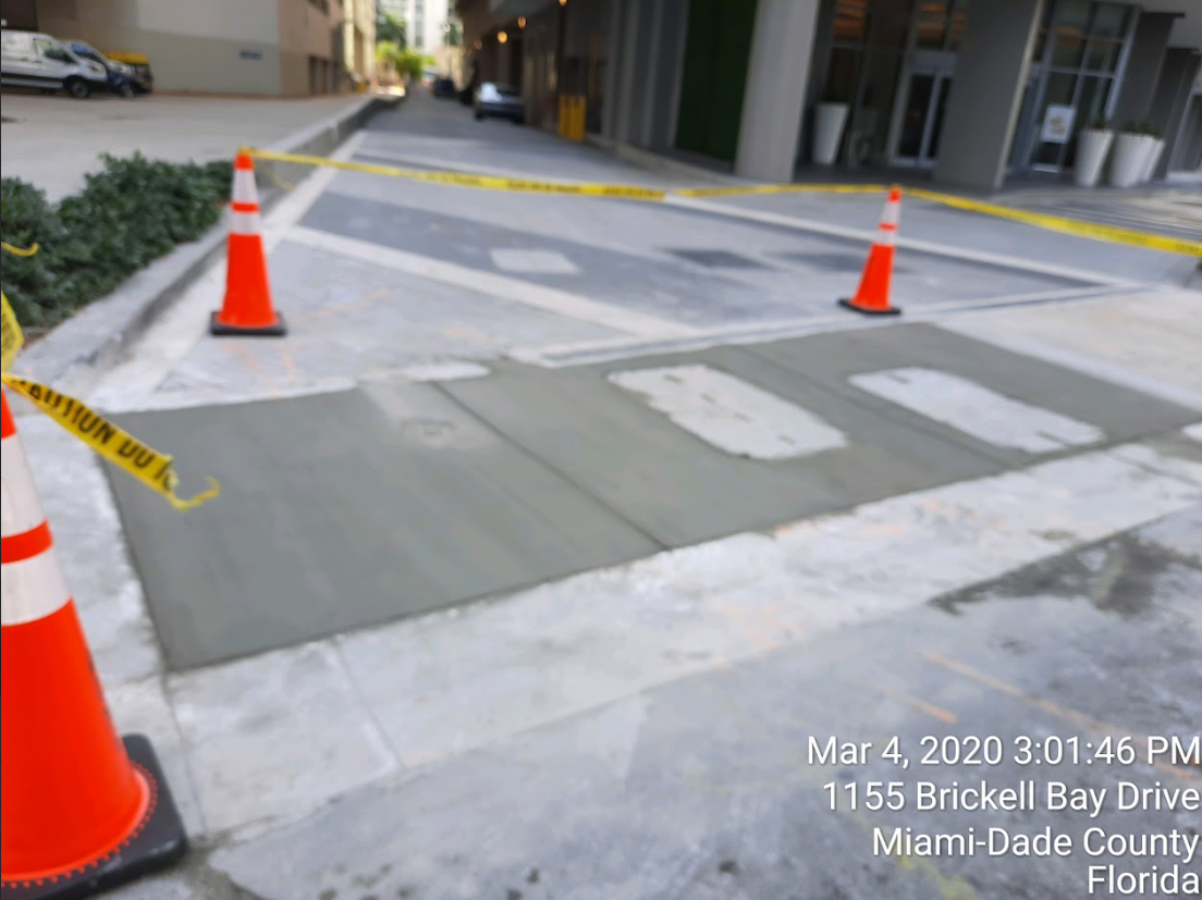 Freshly poured concrete sidewalk section cordoned off with orange traffic cones and yellow caution tape.