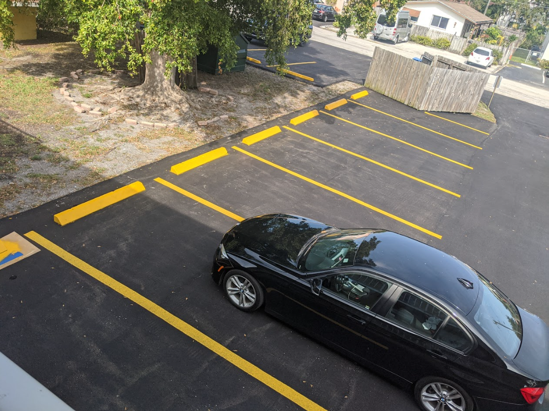 A black sedan parked in an asphalt lot with yellow painted parking stripes and wheel stops next to a grassy area and trees.