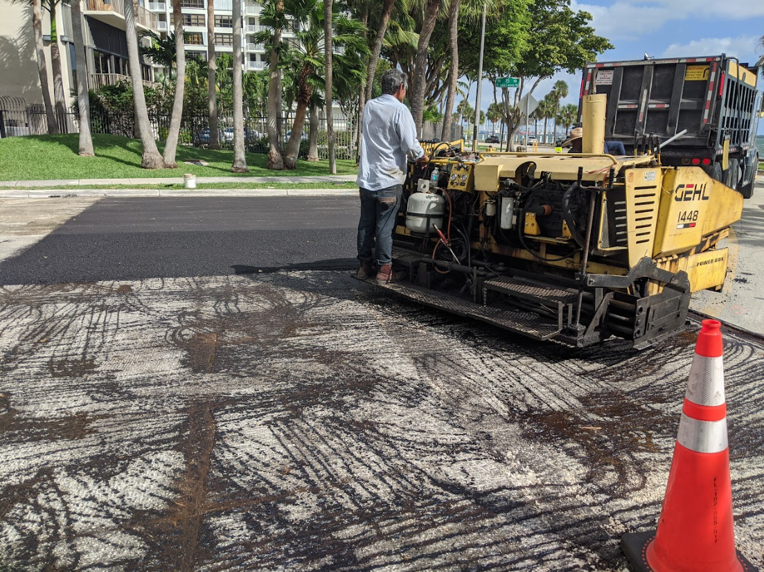 A bright yellow paving machine applies fresh asphalt to a road under a sunny sky near trees and hills.