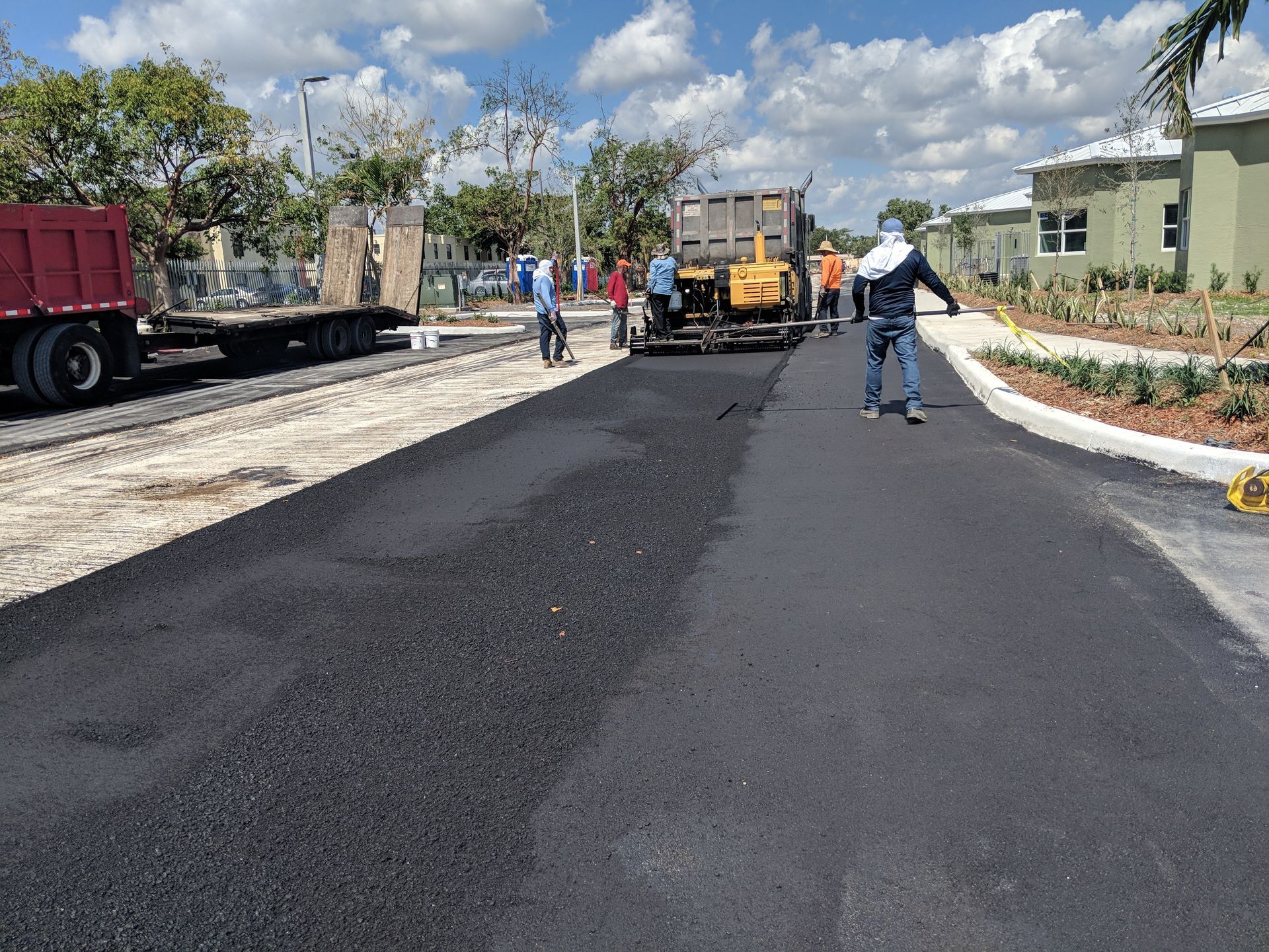 A construction crew paves a residential street with black asphalt under a blue sky with scattered clouds.
