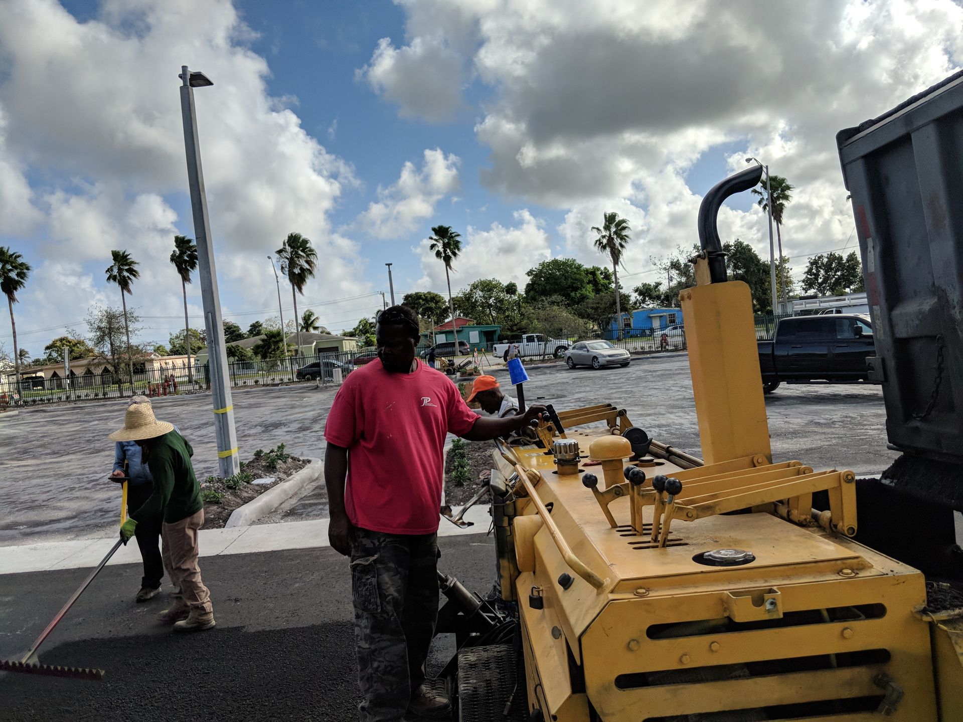 Construction workers perform road paving beside a yellow tractor in an outdoor lot on a sunny day.