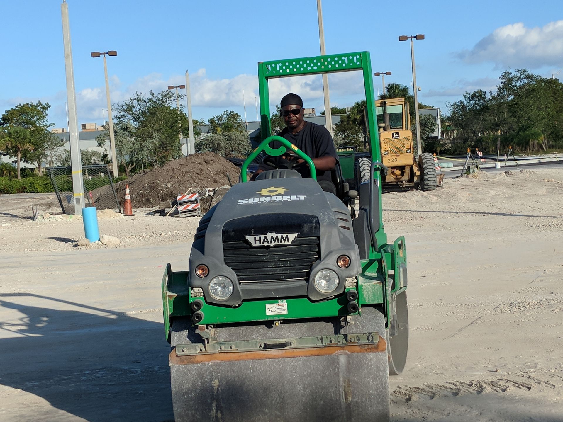 A person operates a green Hamm road roller on a construction site with dirt and heavy machinery in the background.