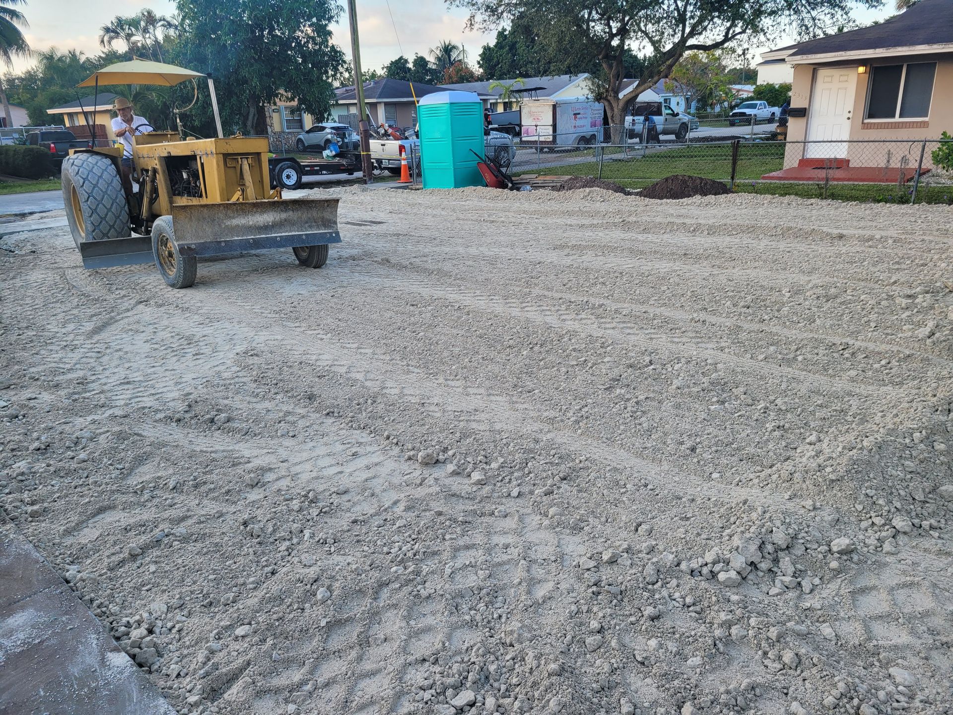 A yellow tractor levels a gravel lot in a residential area, with a portable toilet and houses visible in the background.