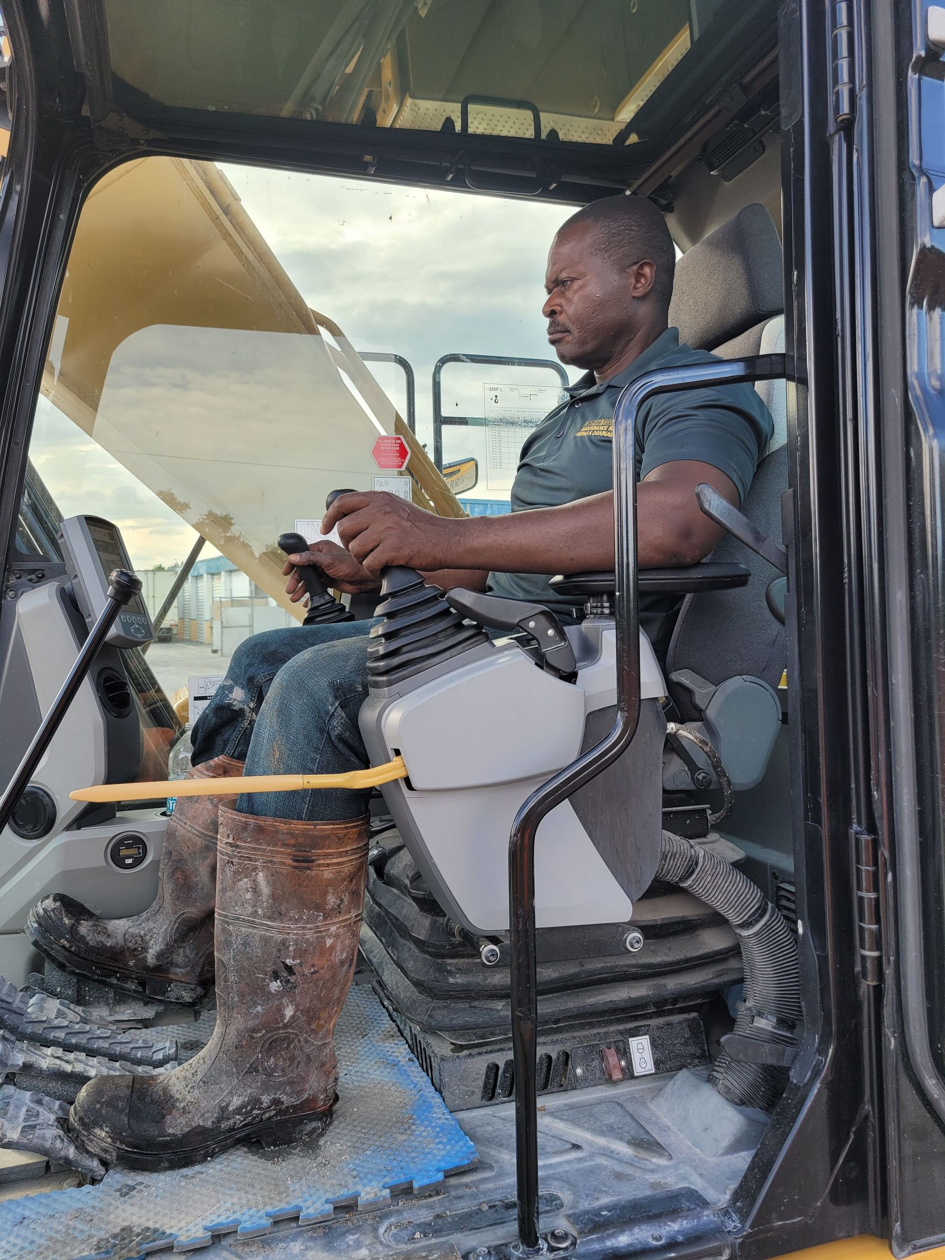 A person sits in the operator's cab of heavy machinery, holding the controls while looking out toward a worksite.