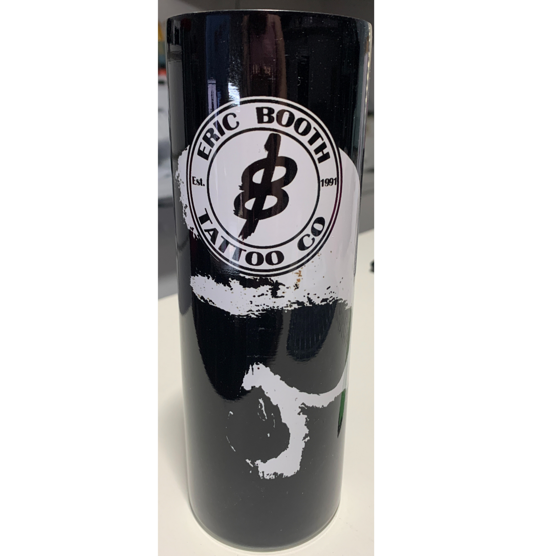 Black cylindrical bottle with a white “Eric Booth Tattoo Co.” logo and abstract white graphic design.