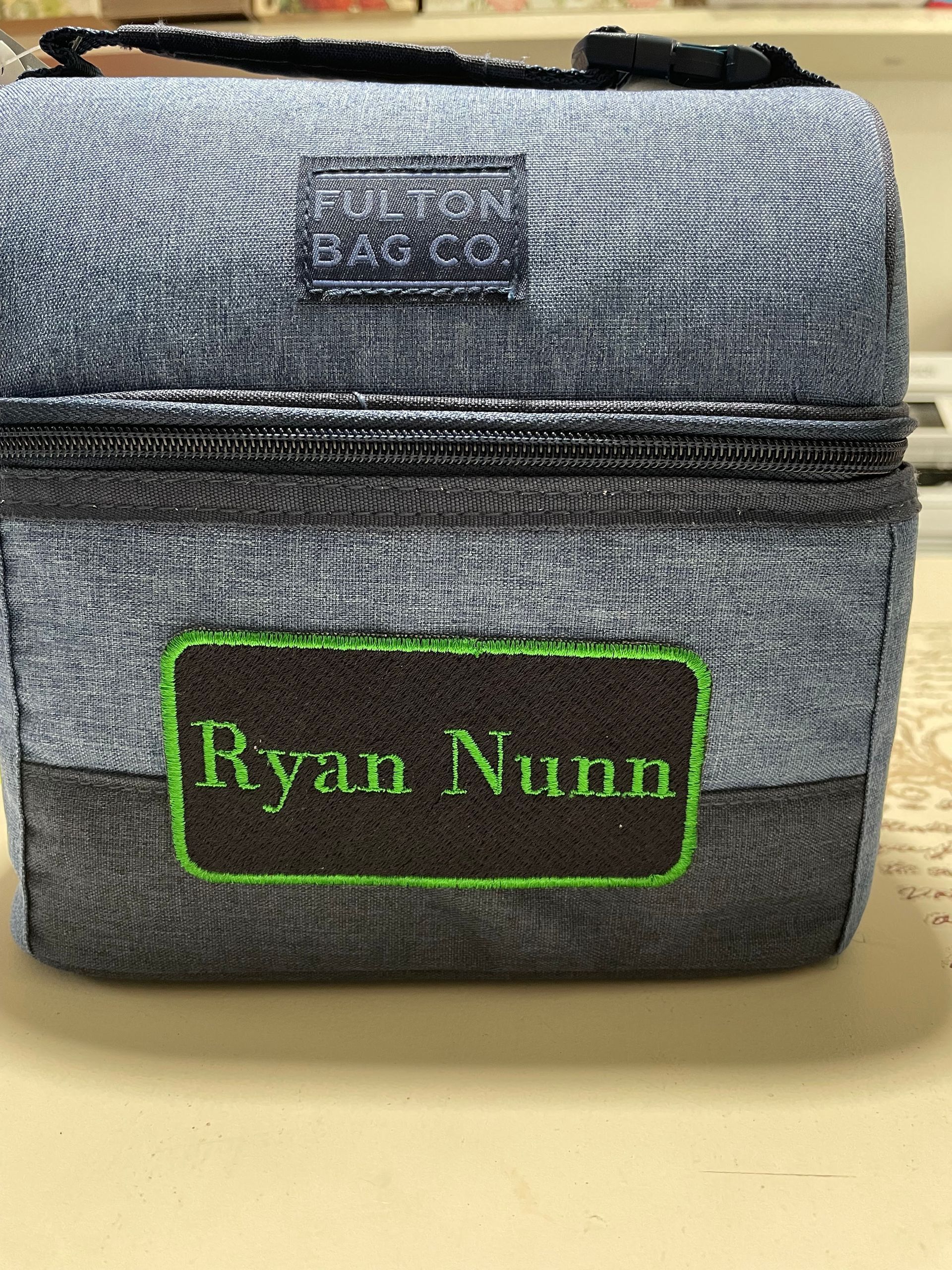 Gray zippered bag with green “Ryan Nunn” name patch on a table