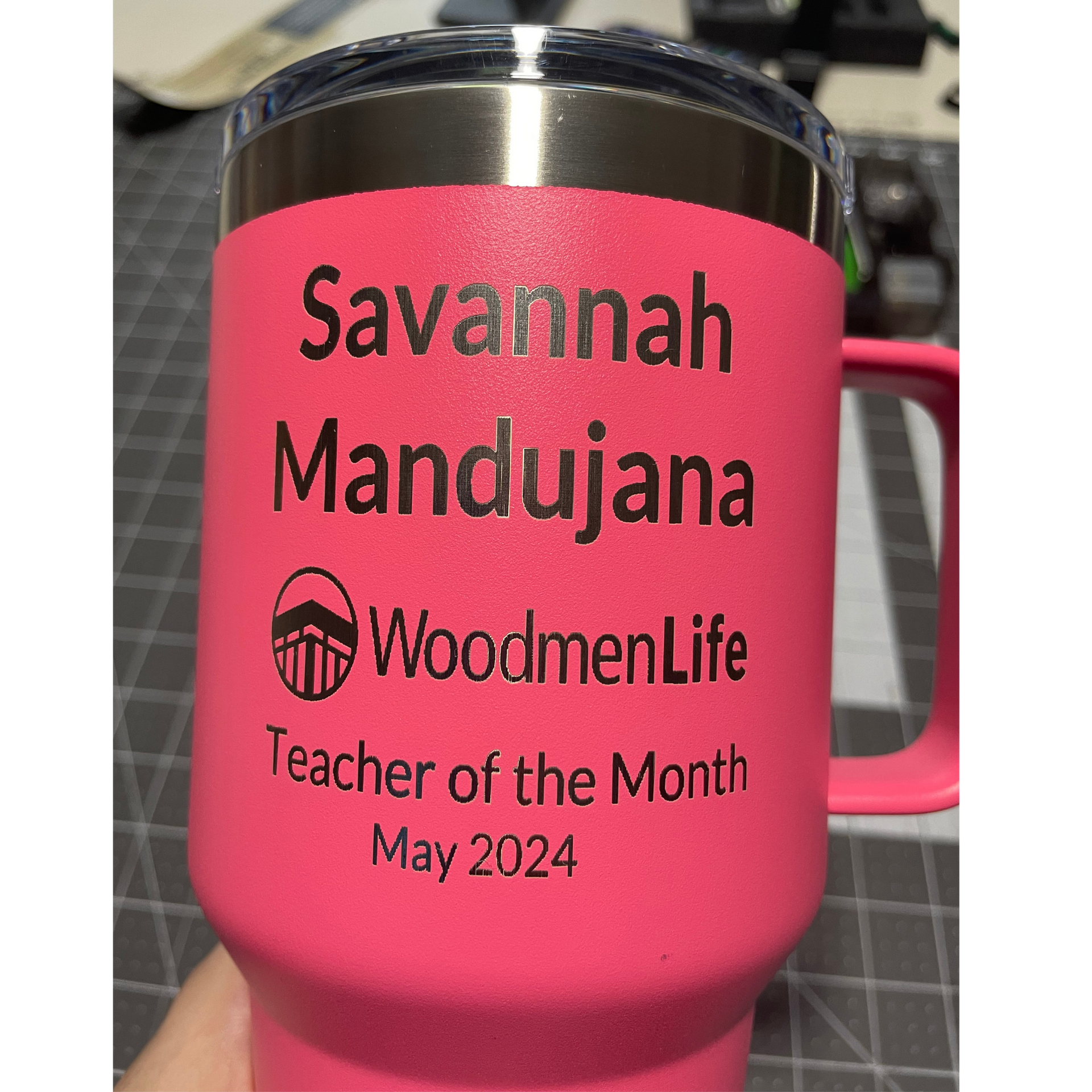 Pink travel mug with black lid and pink handle, labeled “Savannah Mandujana” and “WoodmenLife Teacher of the Month May 2024”