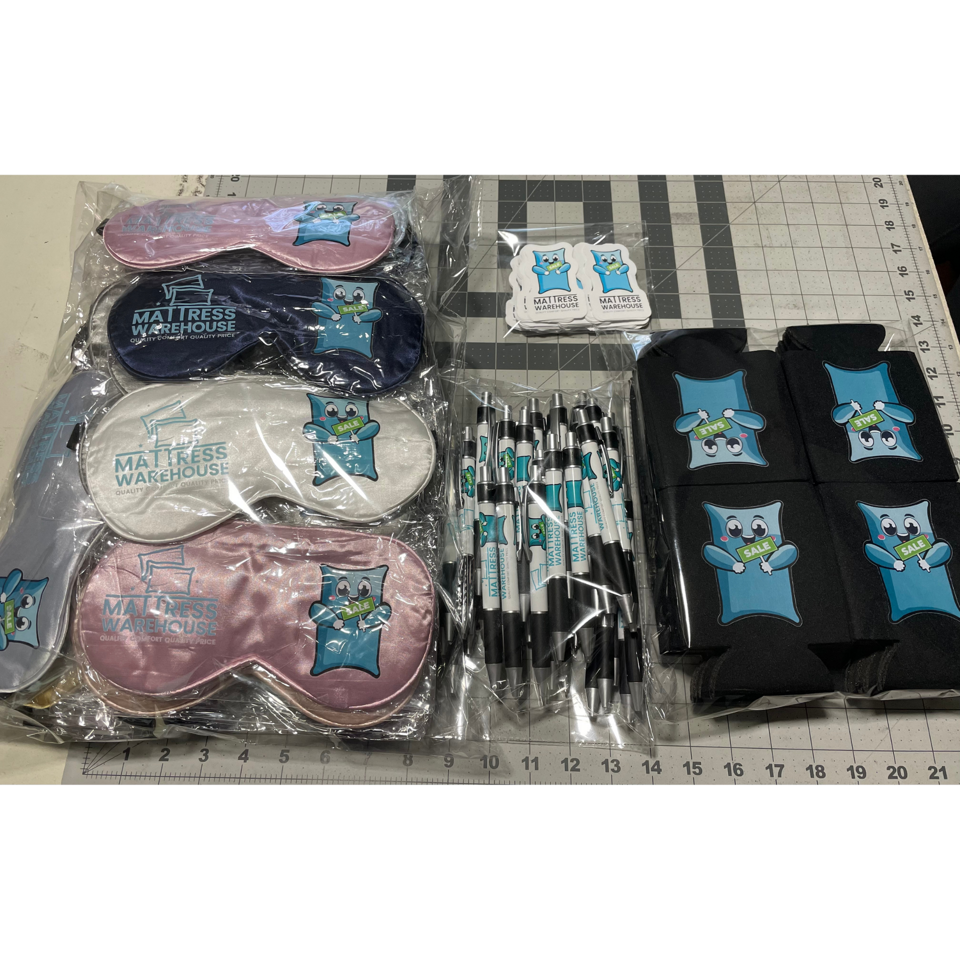Assorted stationery sets on a grid mat: pastel eye masks, pens, stickers, and black pouches with cartoon cat patches