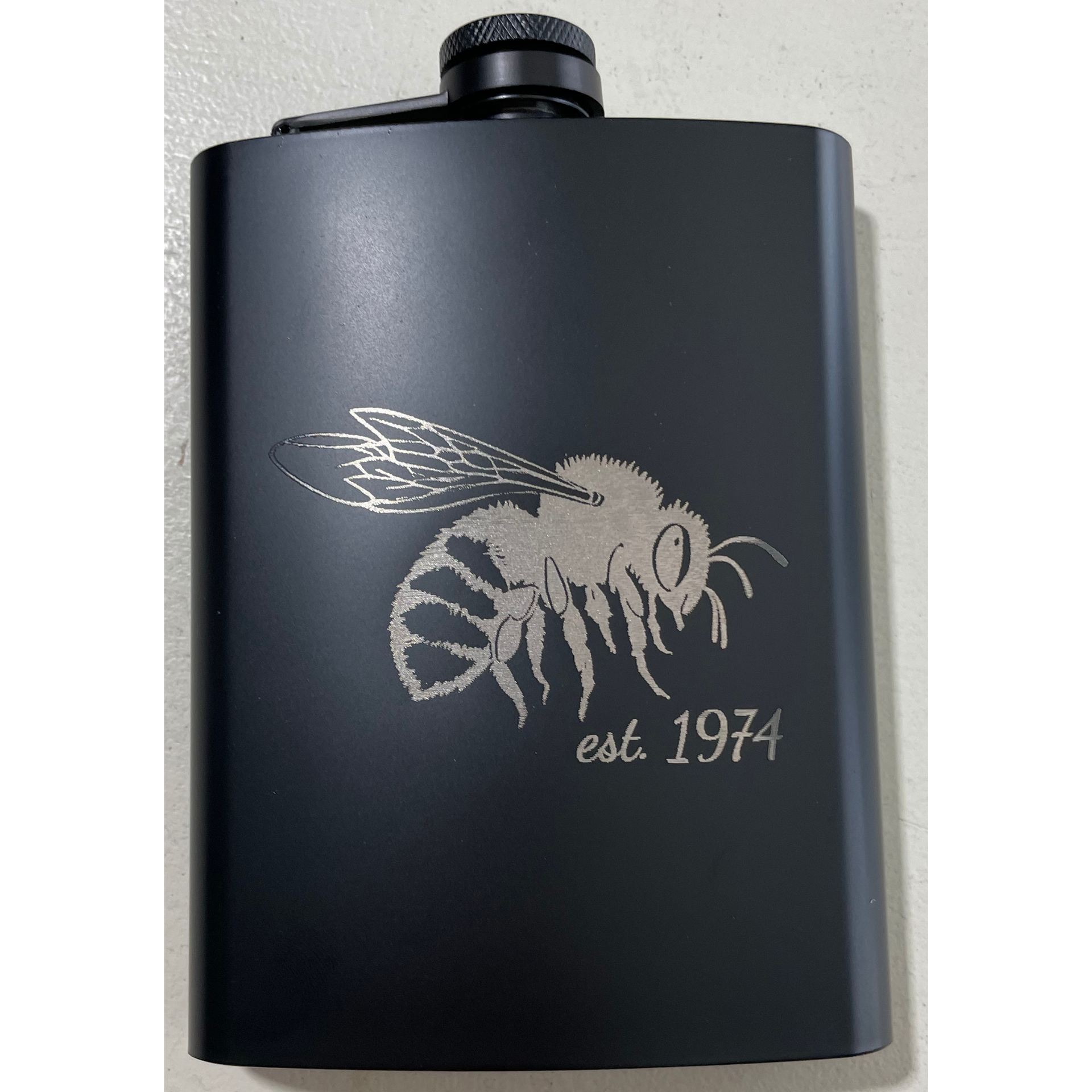 Black metal flask with a silver bee graphic and “est. 1974” text.