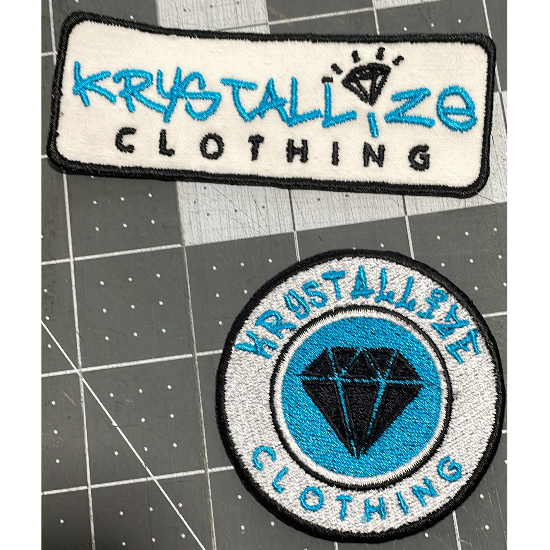 Two Kryztallize Clothing patches, one rectangular and one round, blue and black on gray fabric