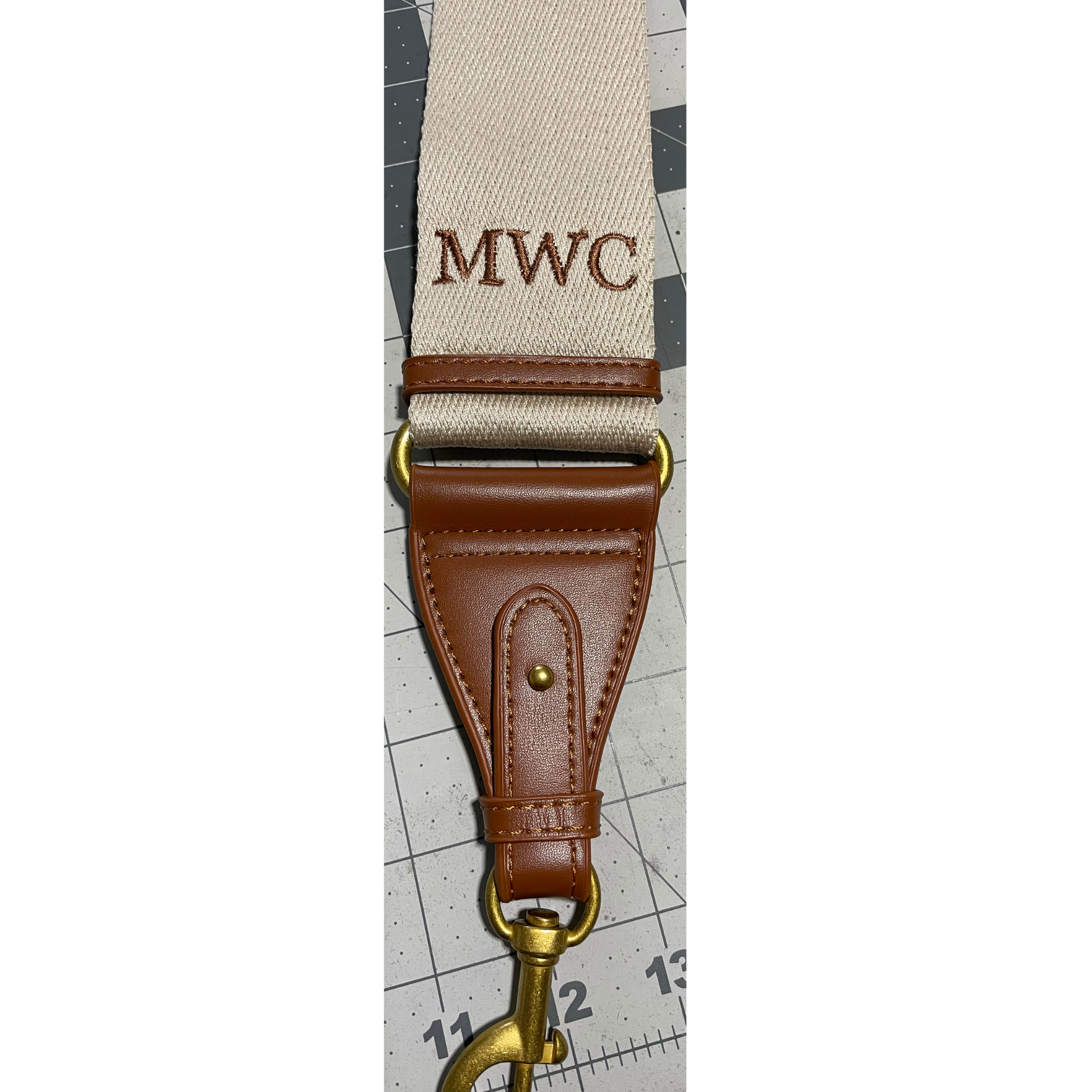 Brown leather camera strap with brass clip and embroidered “MWC” on a gray fabric band