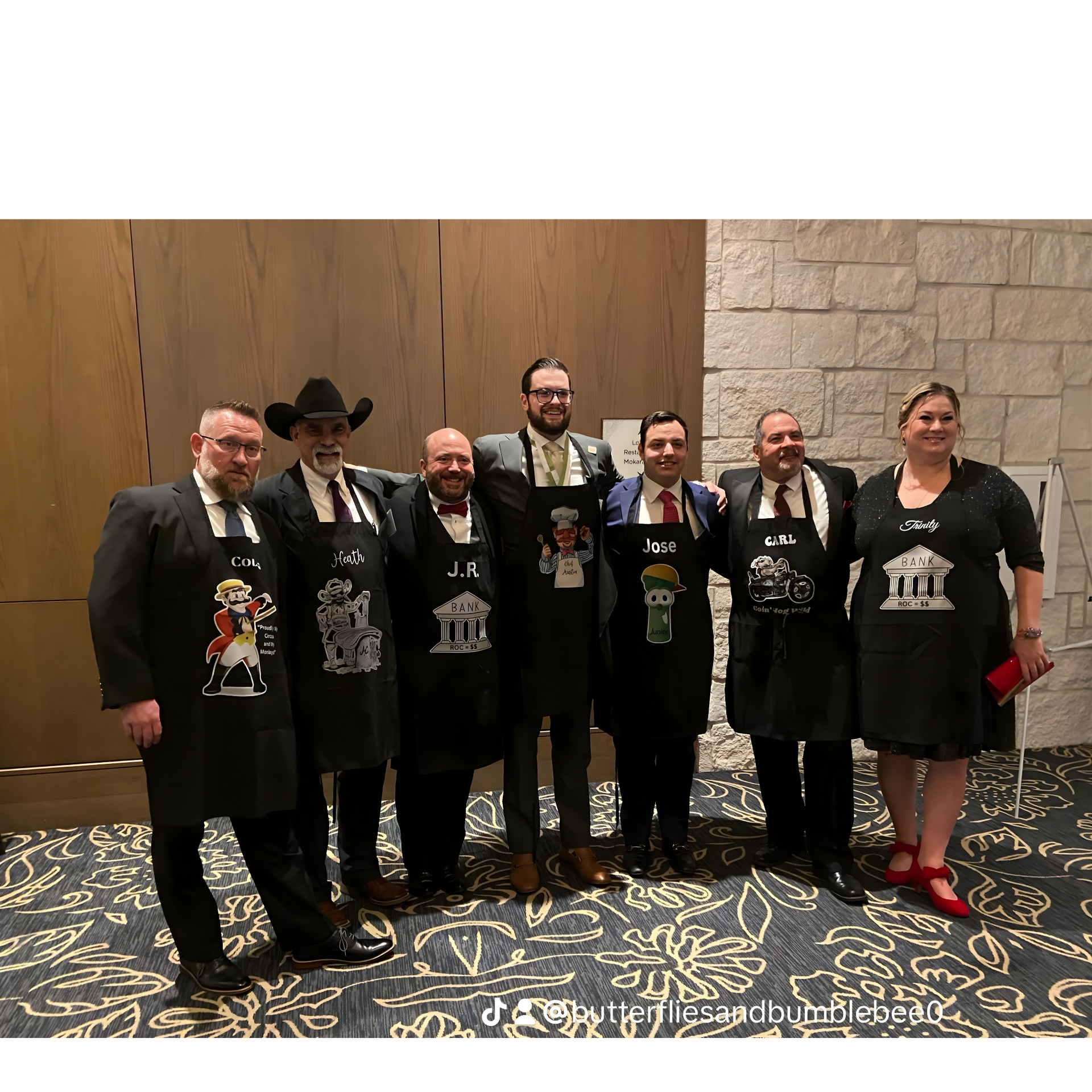 Group of seven people in formal wear with black aprons, posing indoors against a wood and stone wall.
