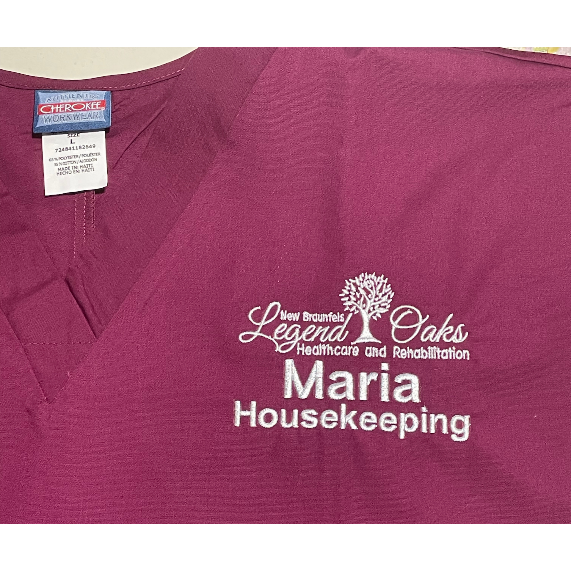 Maroon scrub top with white embroidered name tag reading “Maria Housekeeping” and Legends Oaks logo.
