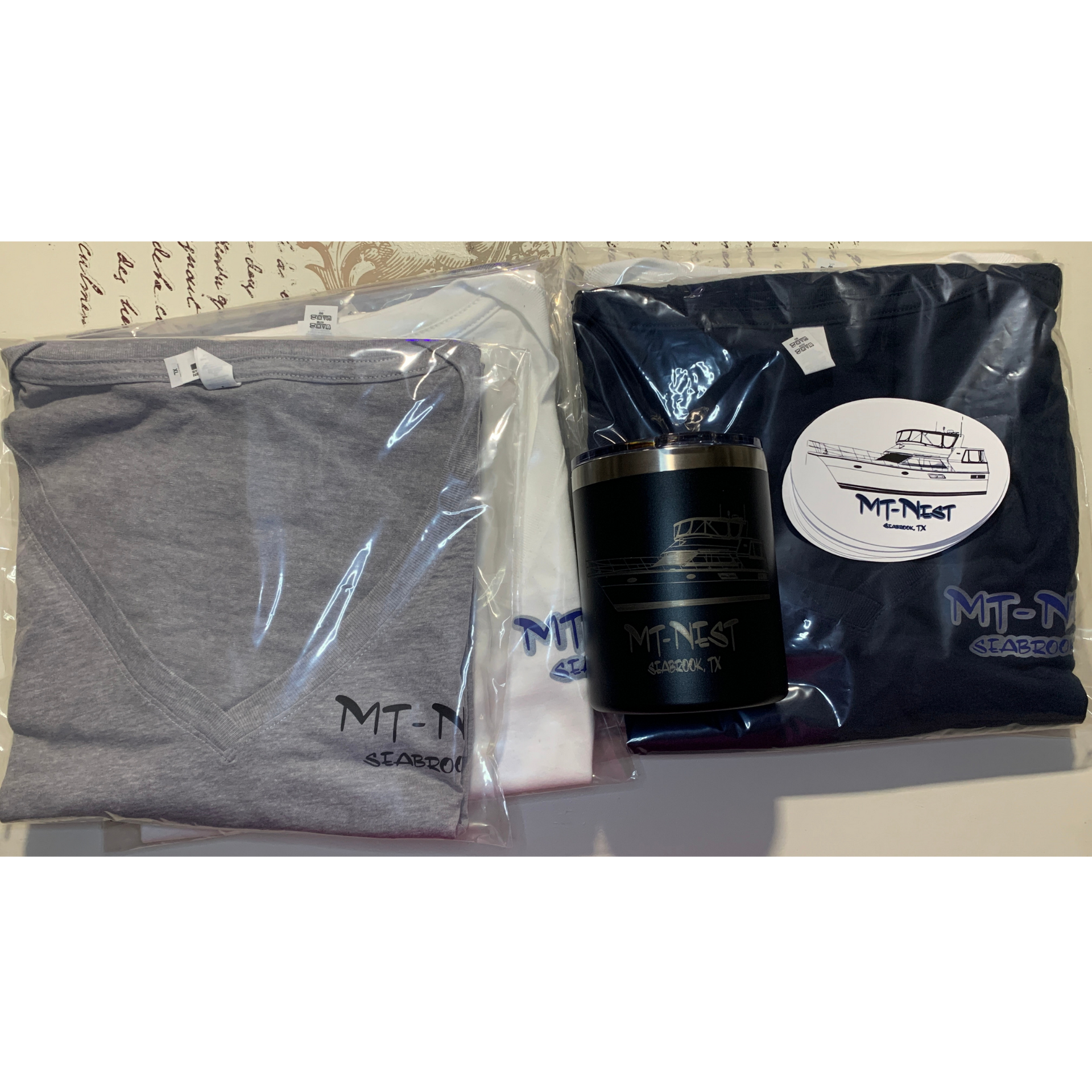 Folded gray and navy MTM apparel in plastic, with a black tumbler and white product tag on a table.