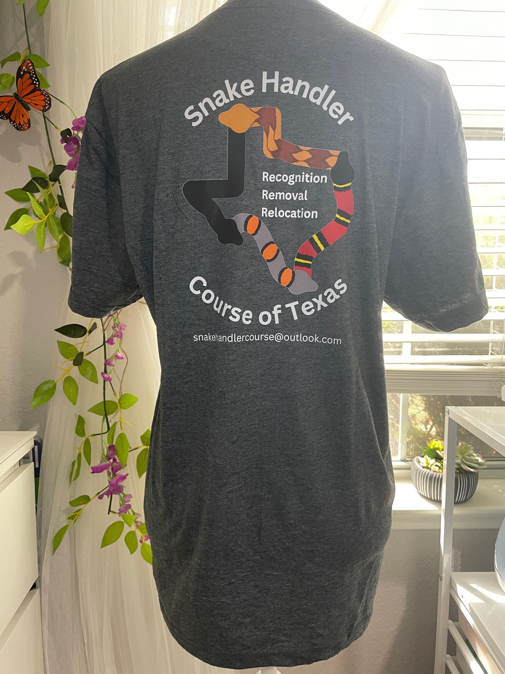 Black T-shirt with a snake graphic and text reading “Snake Handler” and “Course of Texas,” hanging by a window.