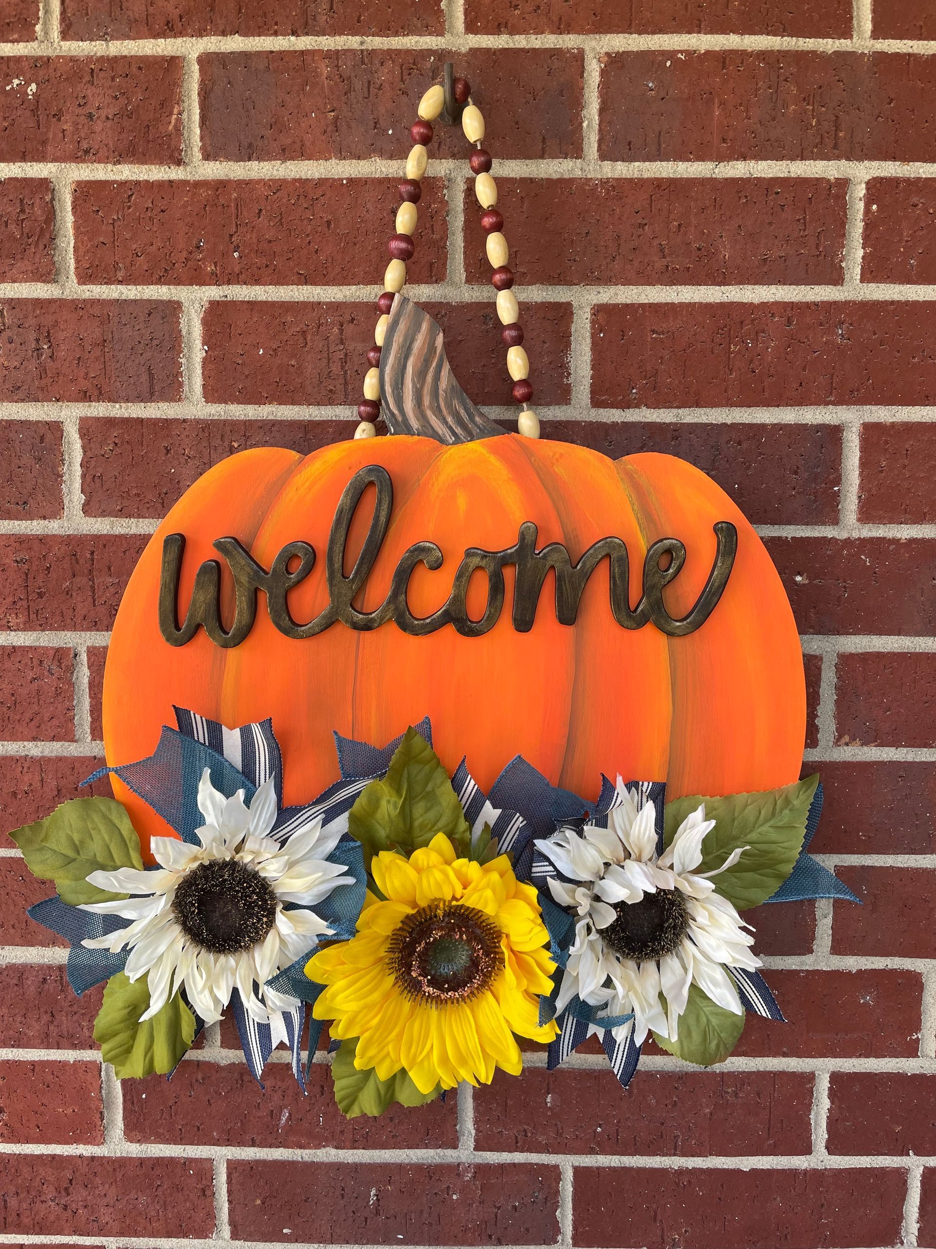 Orange “welcome” pumpkin wreath with sunflower and flower accents on a brick wall