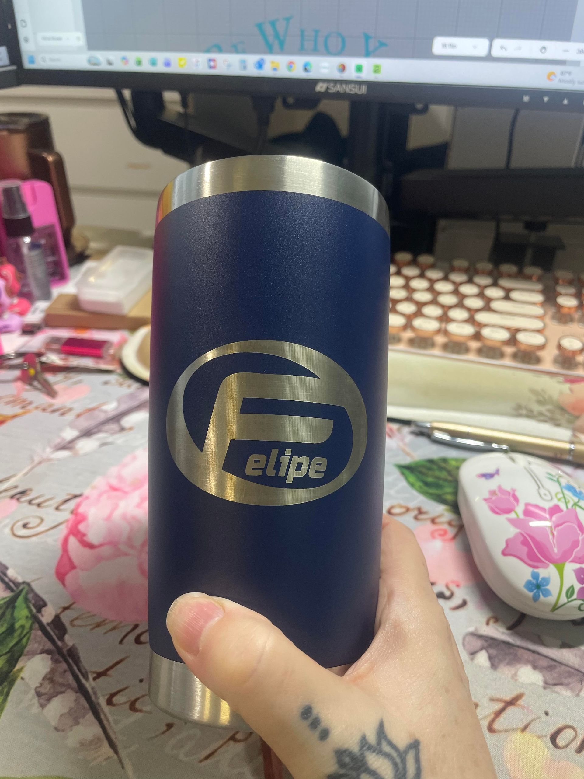 Hand holding a blue E1lbe stainless steel tumbler in front of a desk with a monitor and makeup items.