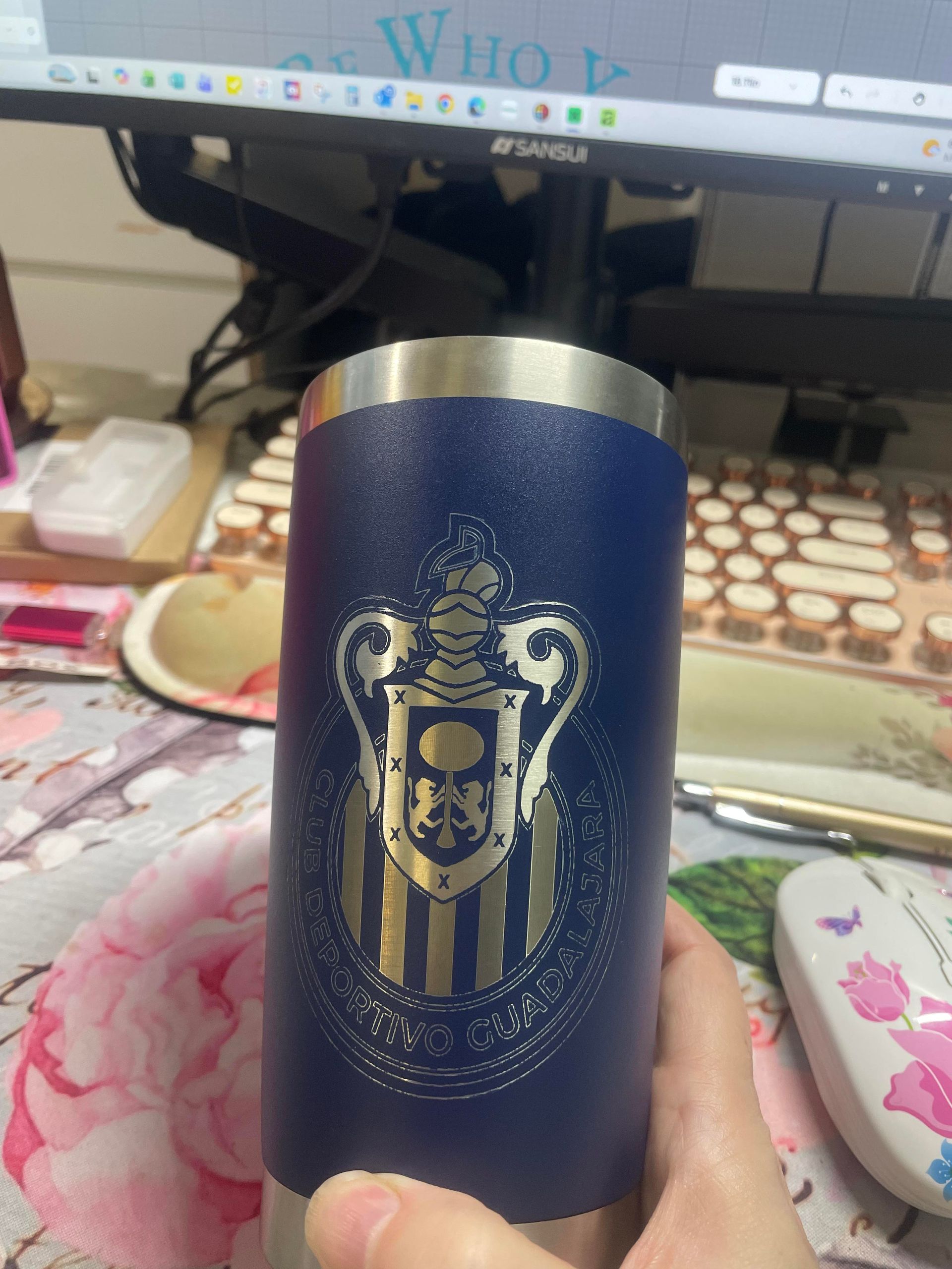 Blue insulated tumbler with a gold crest logo on a desk in front of a computer monitor.