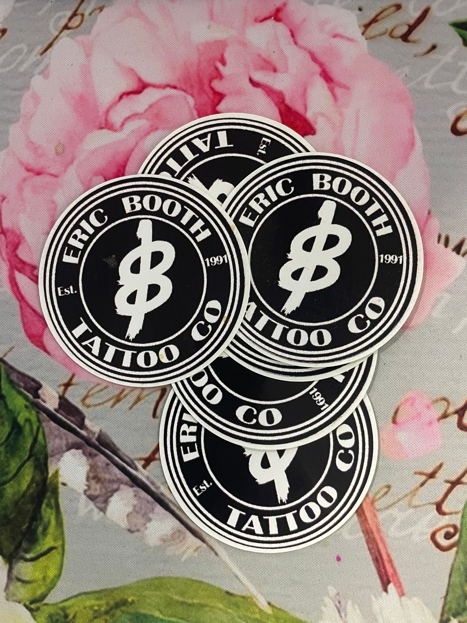 Three black-and-white circular tattoo shop stickers on a pink floral background