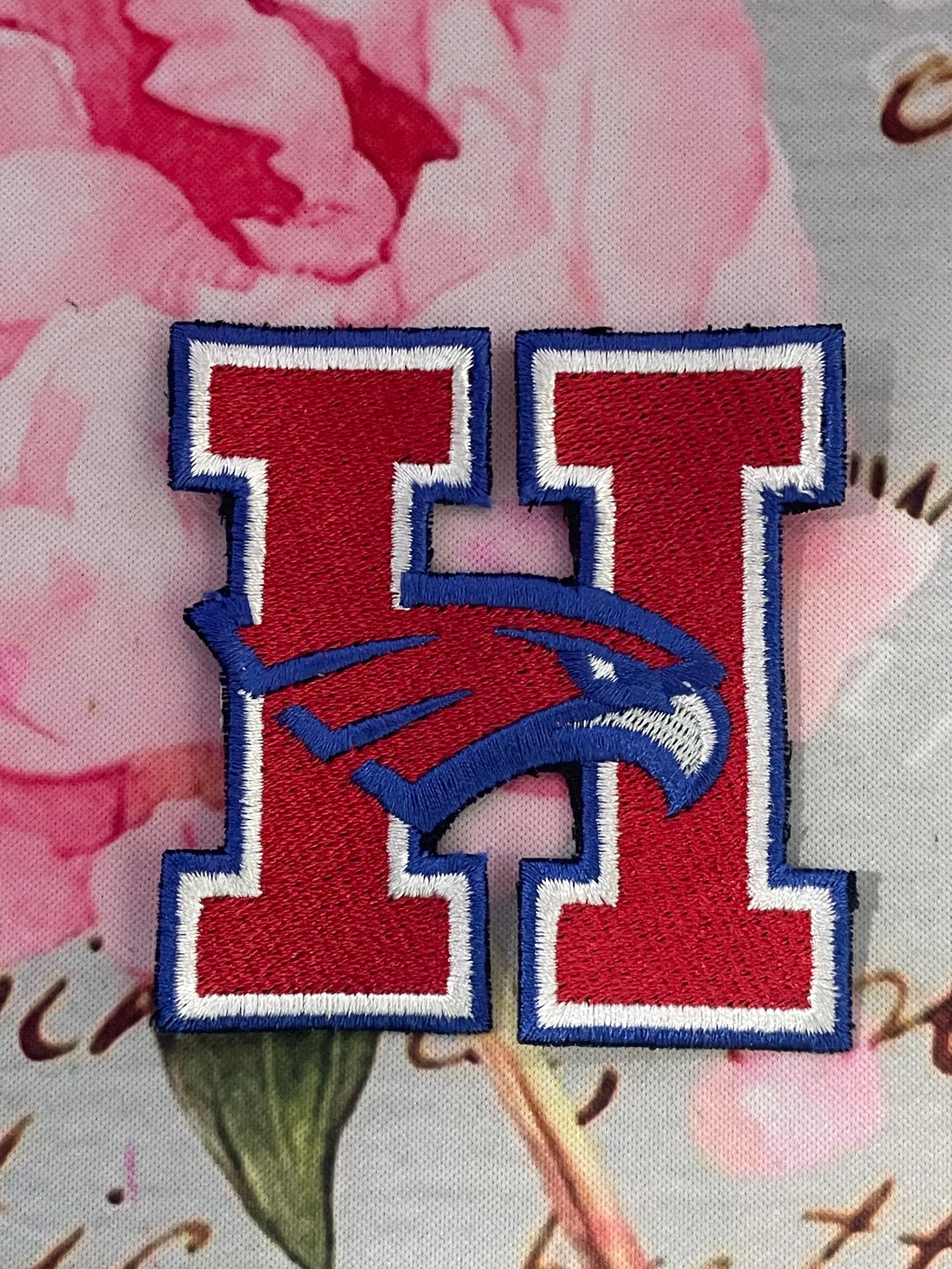 Red and blue H logo with a bird head, embroidered on pink floral fabric.