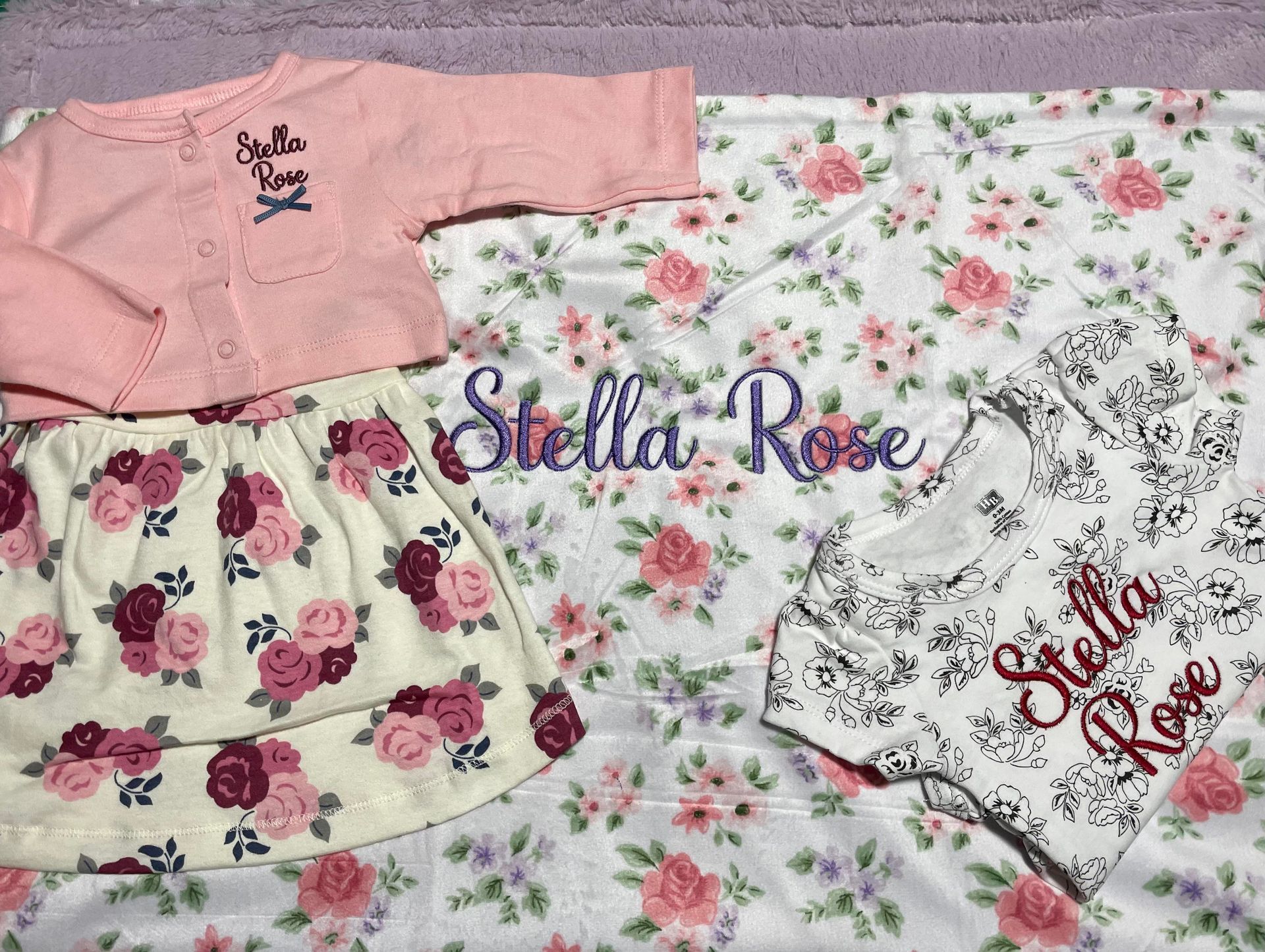 Pink and floral baby outfits with “Stella Rose” embroidery laid out on a white surface