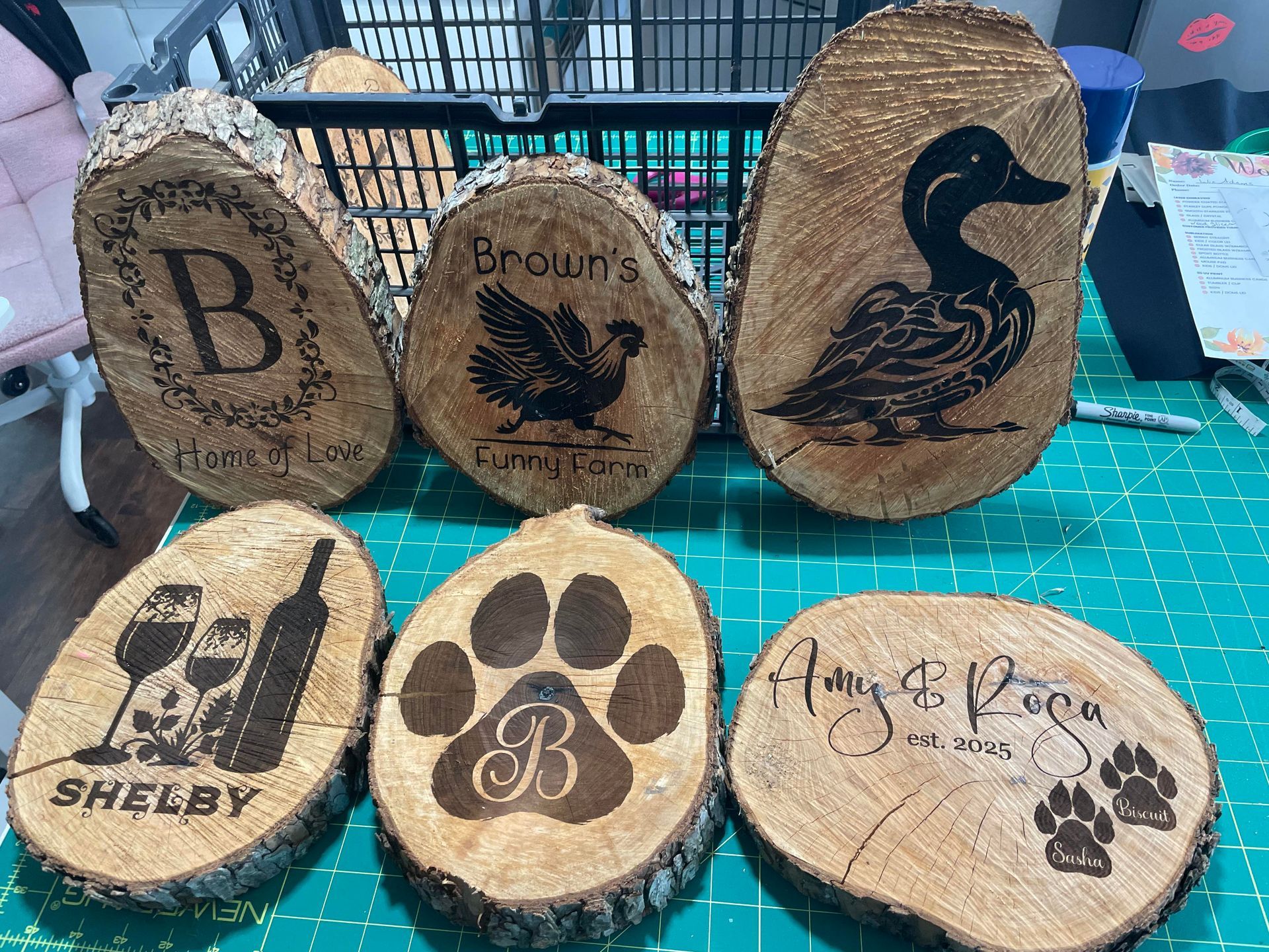 Six decorated oval wood slices with engraved pet and monogram designs on a green cutting mat.
