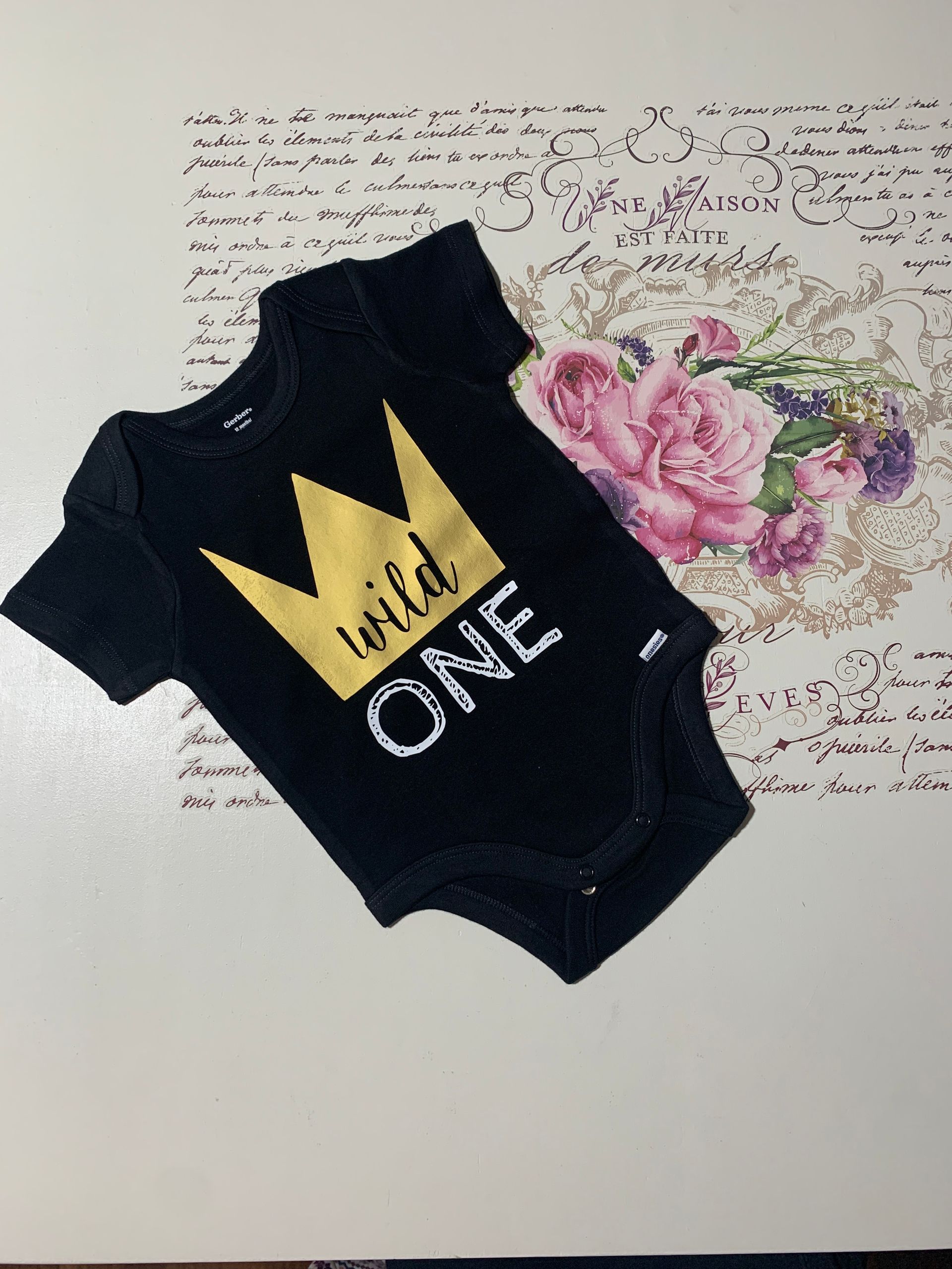 Black baby onesie with gold crown and “Wild ONE” text, beside pink flowers on a handwritten background