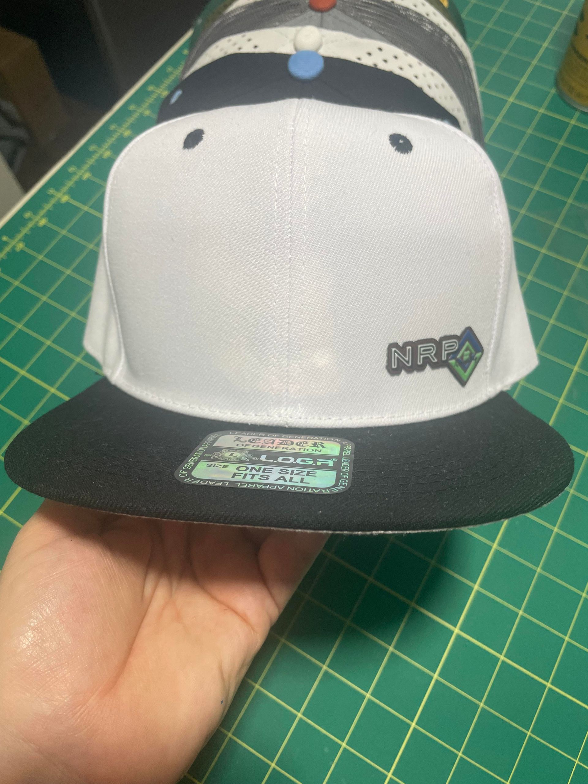 White and black baseball cap with cartoon face and small logo, held over a green cutting mat.