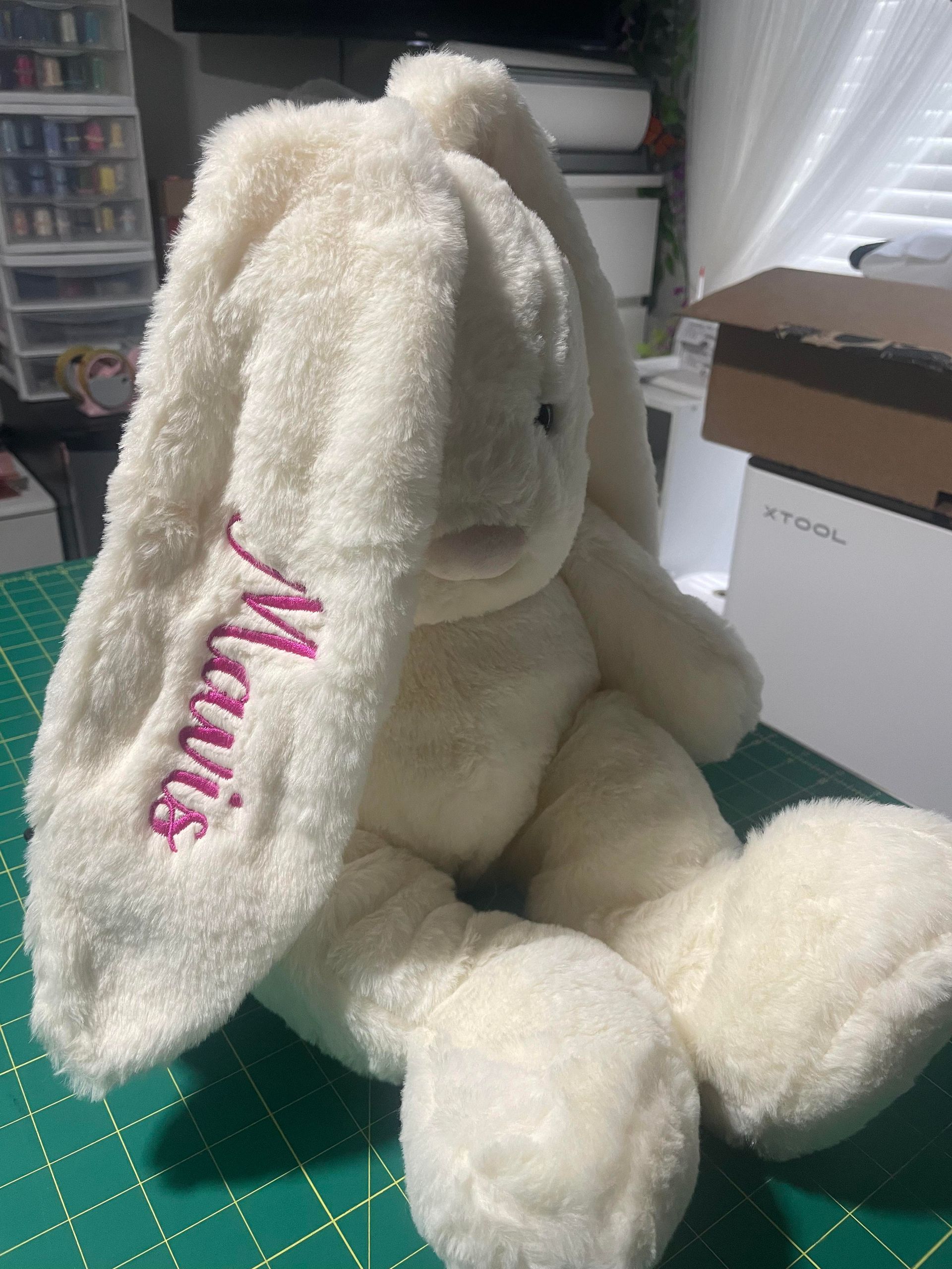 Cream plush bunny with pink “Mimi” embroidery, sitting on a cutting mat in a craft room.