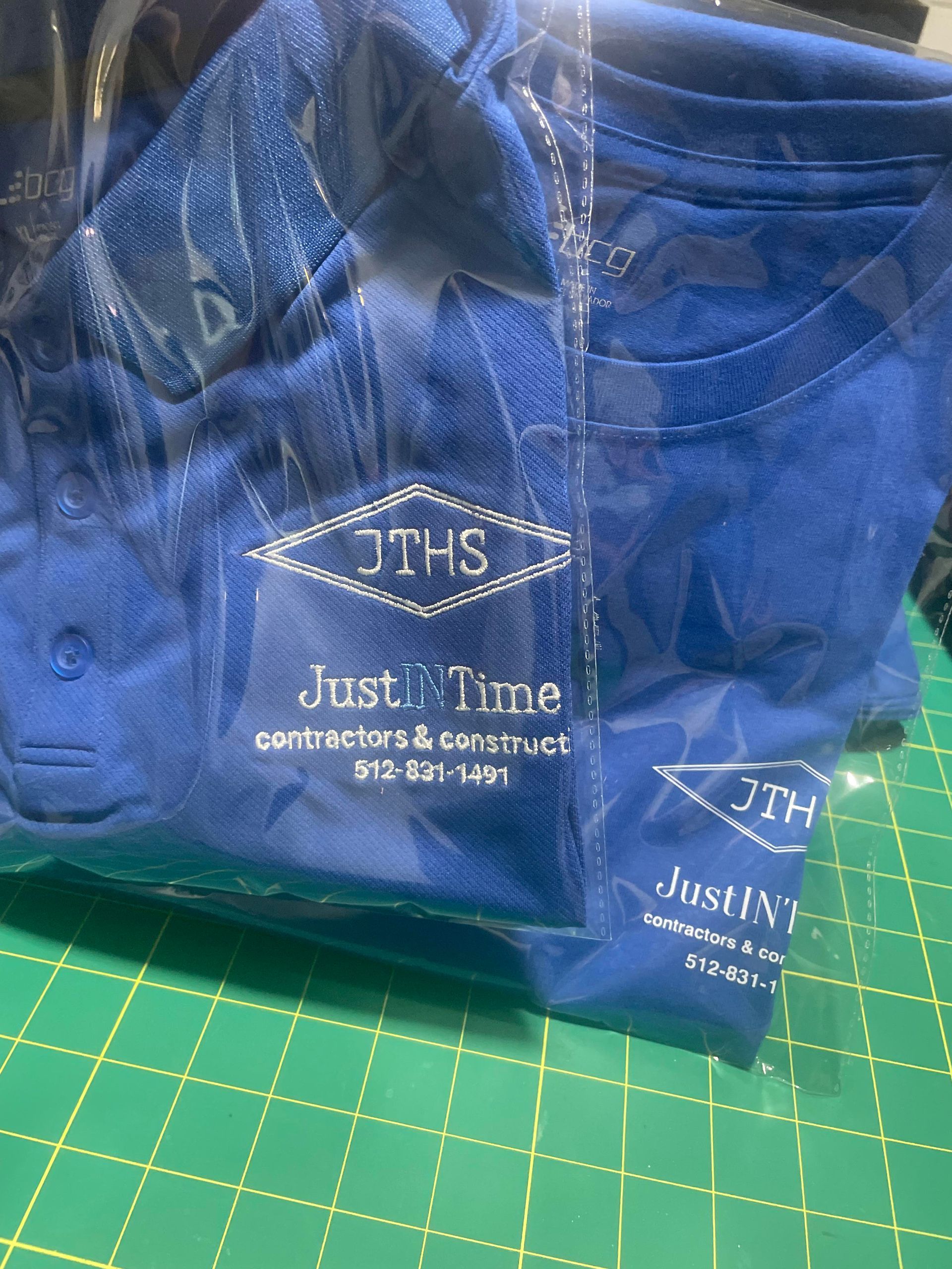 Blue T-shirts in clear plastic packaging on a green cutting mat, with “Just J Time” logo and text.