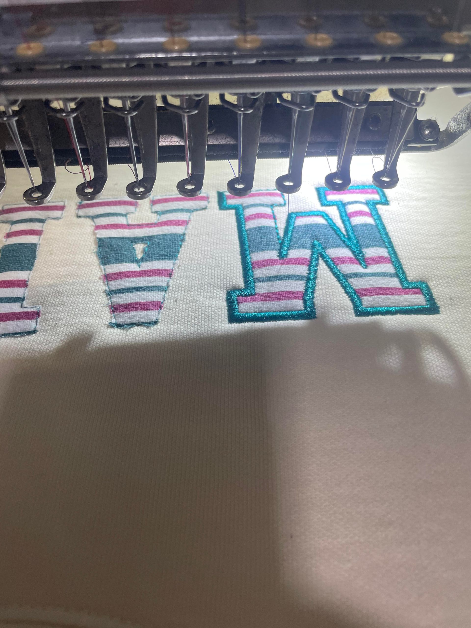 Embroidery machine stitching colorful striped letters on white fabric, spelling “V” and “W”