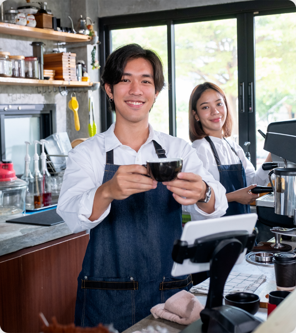 Barista smiling, holding coffee cup; another person in background at counter. Cafe setting.