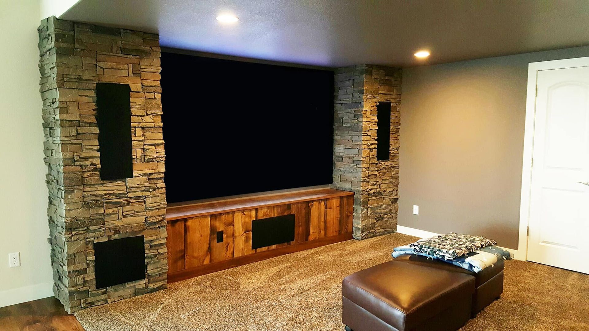 Modern living room with large wall-mounted TV, stone fireplace, carpet, and brown ottoman.