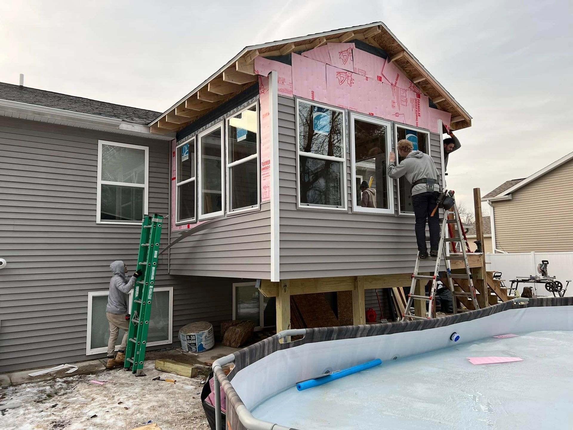 Workers renovating the side of a house, with exposed insulation and a ladder beside an empty above-ground pool.