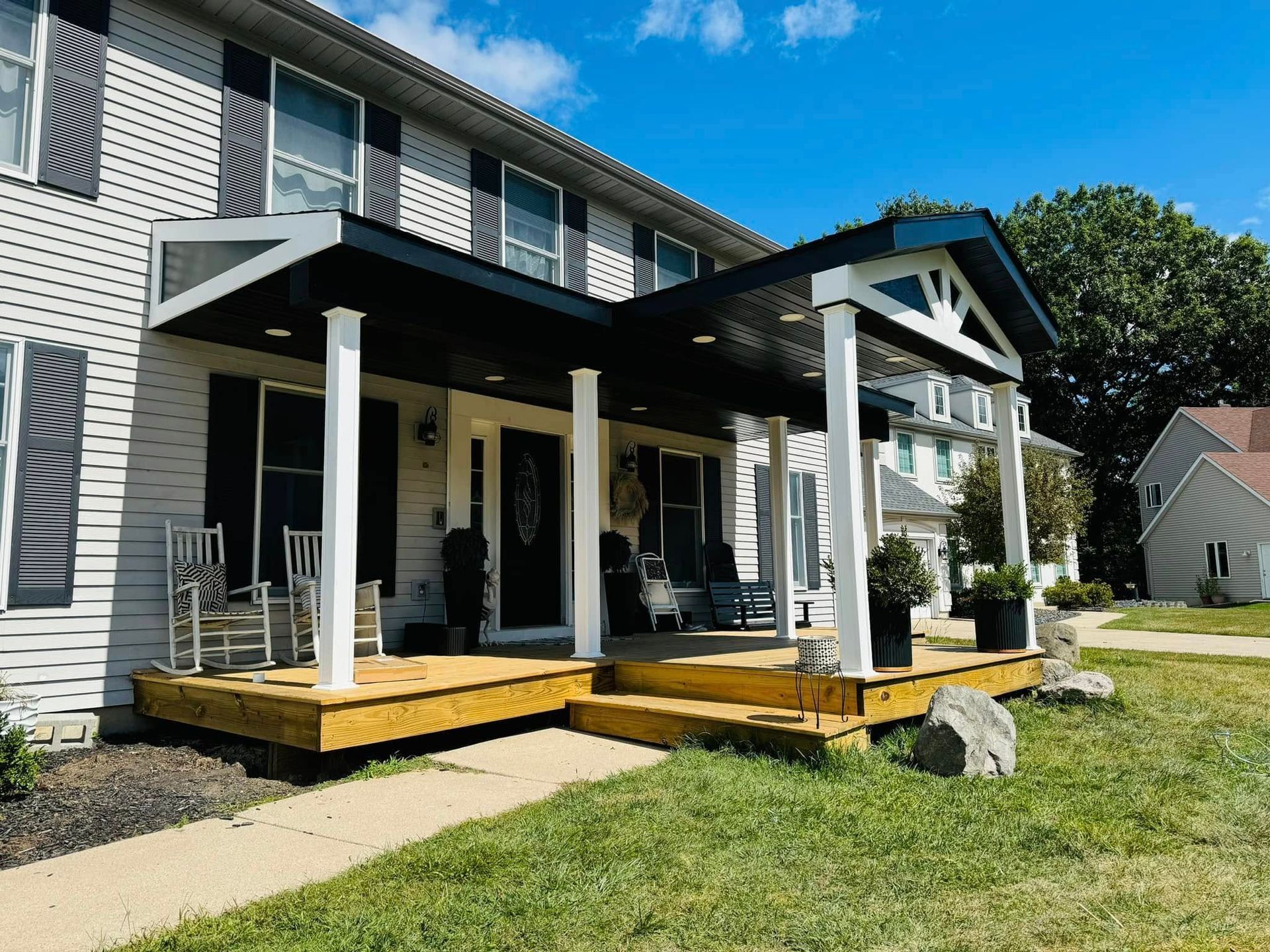 Two-story house with a front porch, wooden steps, and black roof under a blue sky