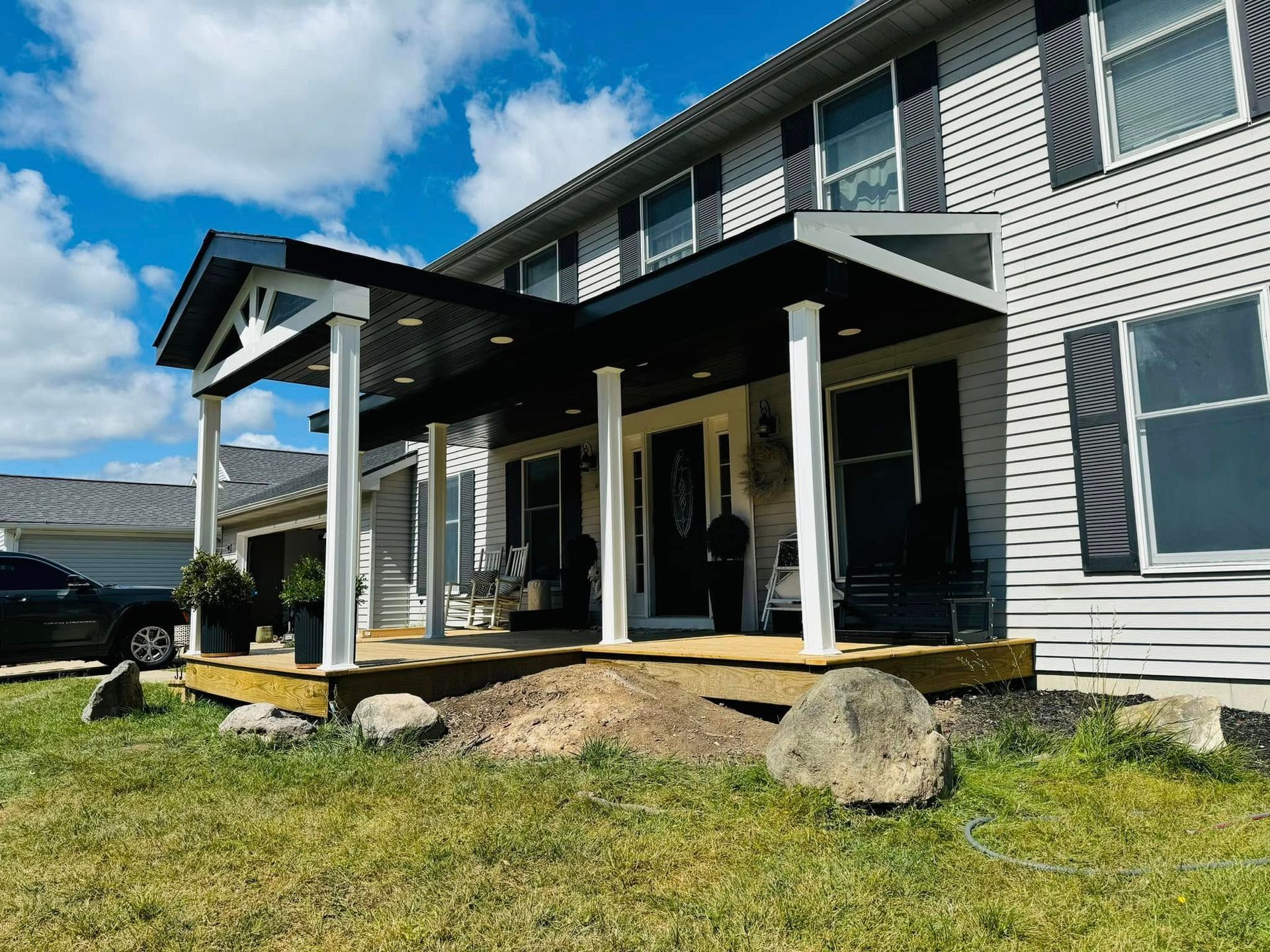 Two-story gray house with a covered porch, black shutters, and a grassy yard with large rocks