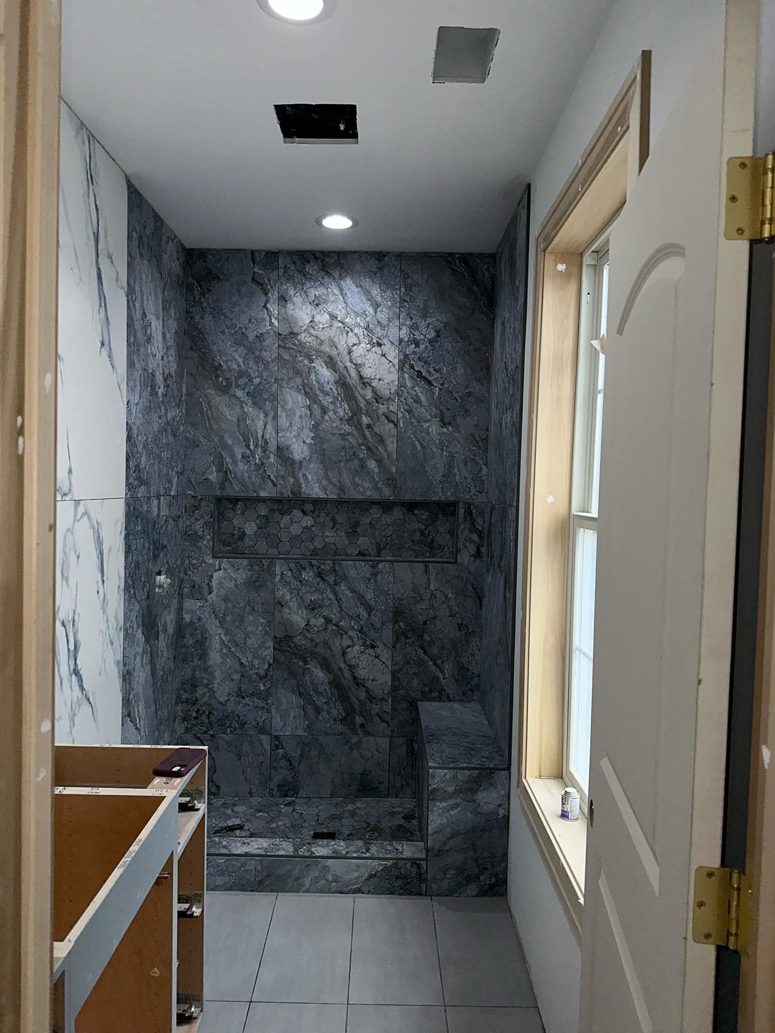 Narrow bathroom with marble shower wall, white tiled floor, window, and vanity under recessed lights