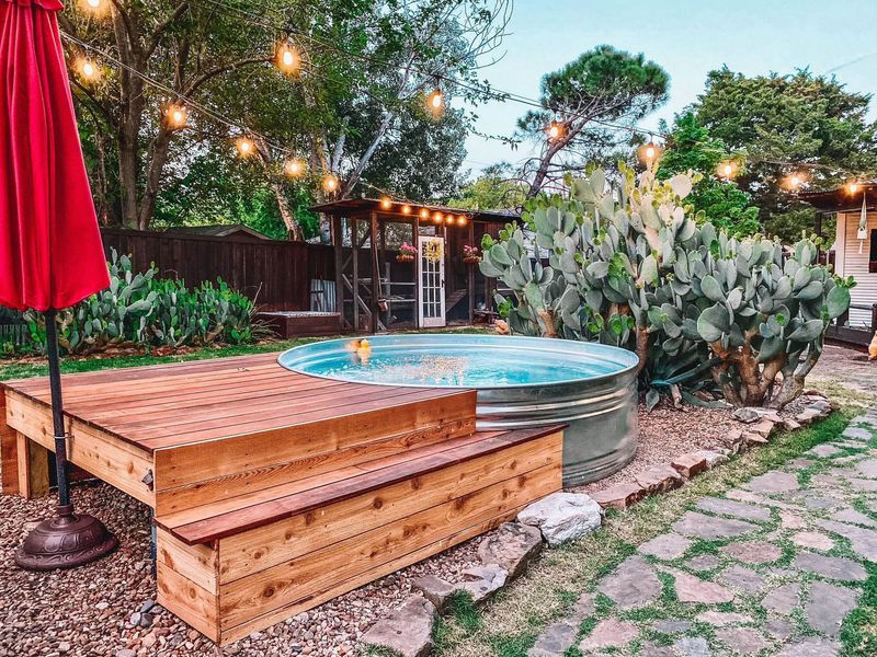 Wooden hot tub on a stone patio in a leafy backyard with string lights and a red umbrella
